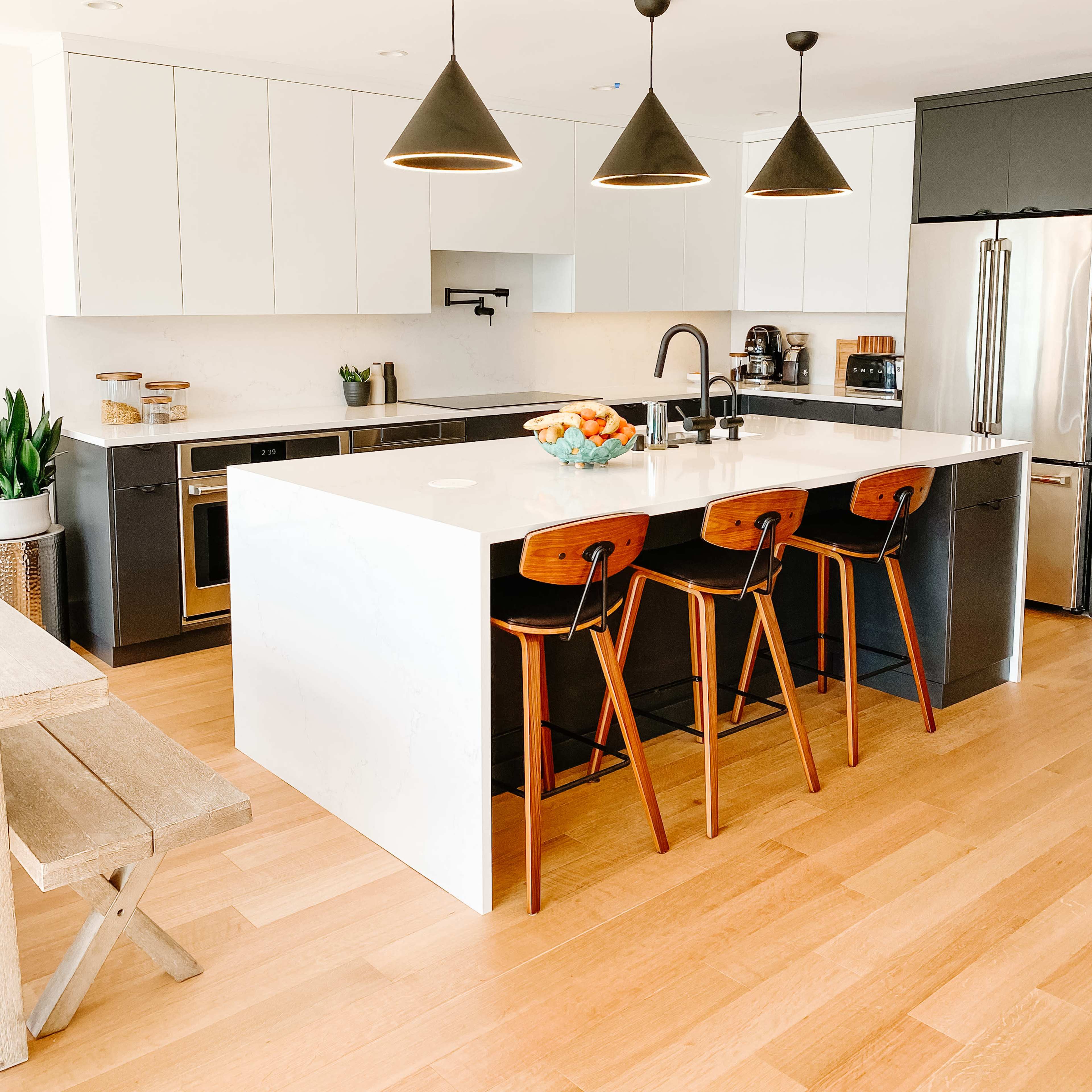 A modern kitchen features a large white island with three wooden bar stools, sleek cabinetry, and pendant lights above.