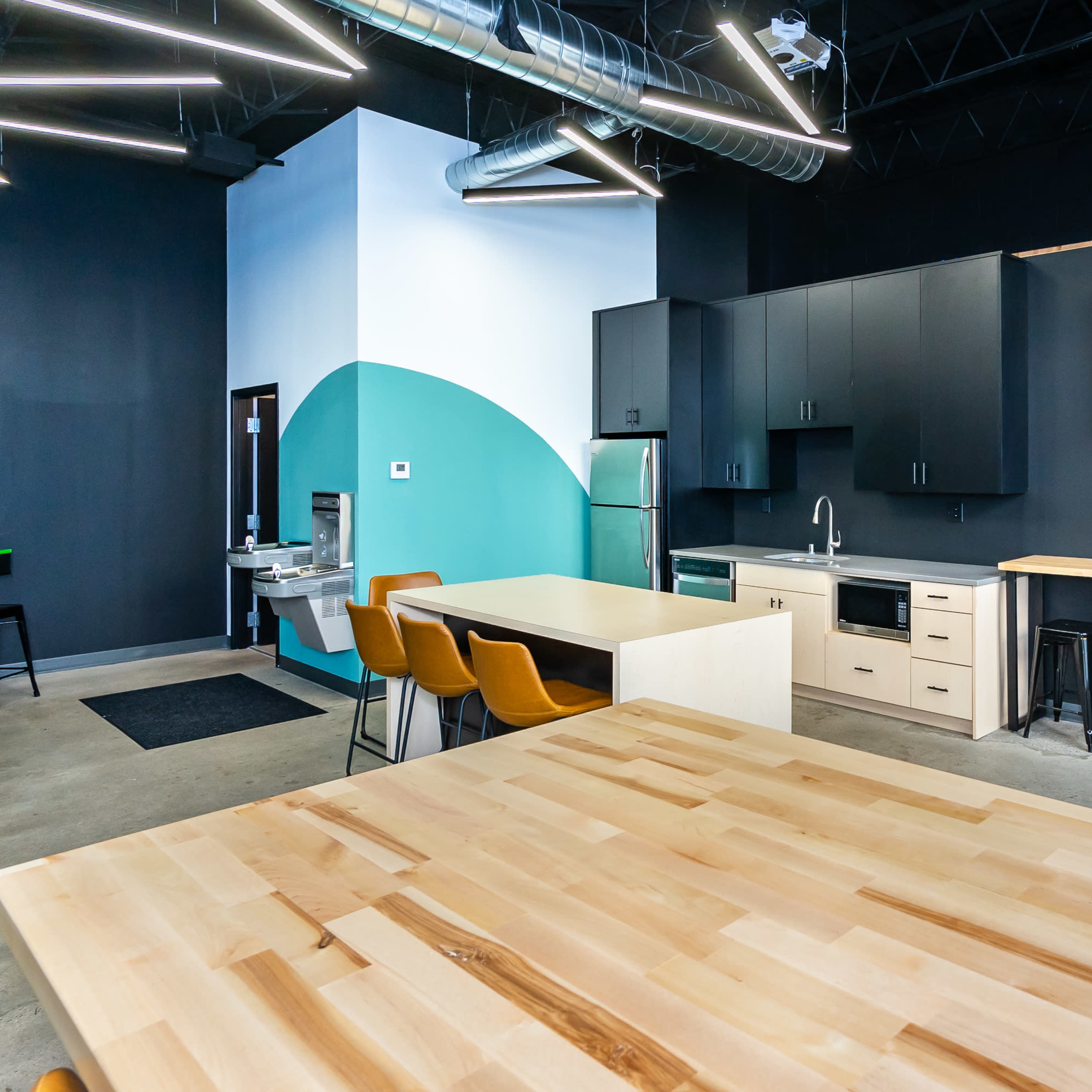 A modern kitchen and dining area with a large wooden table, sleek black cabinetry, and a wall-mounted TV displaying content.