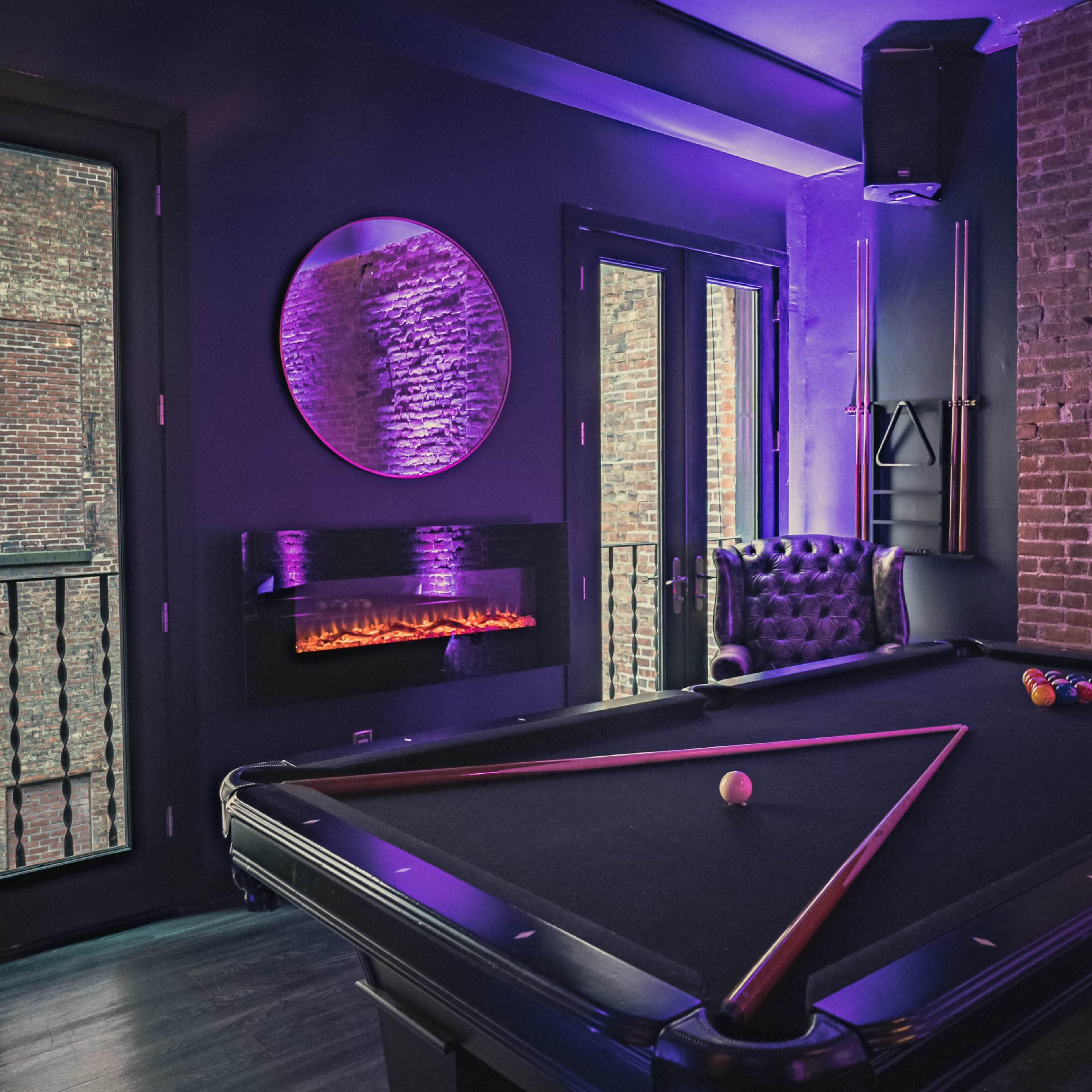 A billiard table is positioned in a dimly lit room with exposed brick walls and a modern electric fireplace.