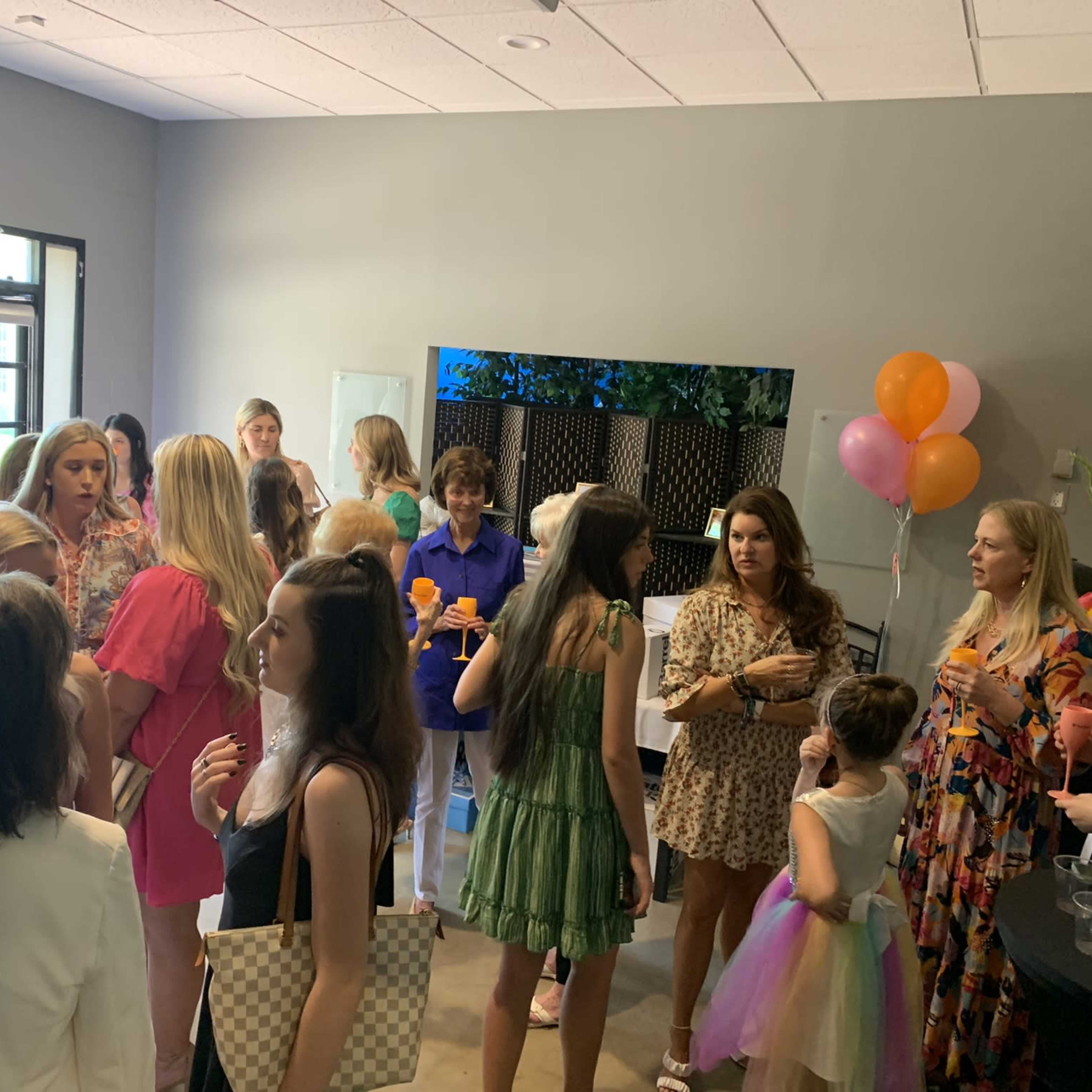 A group of women and children socialize at a gathering inside a bright room, with balloons and drinks visible.