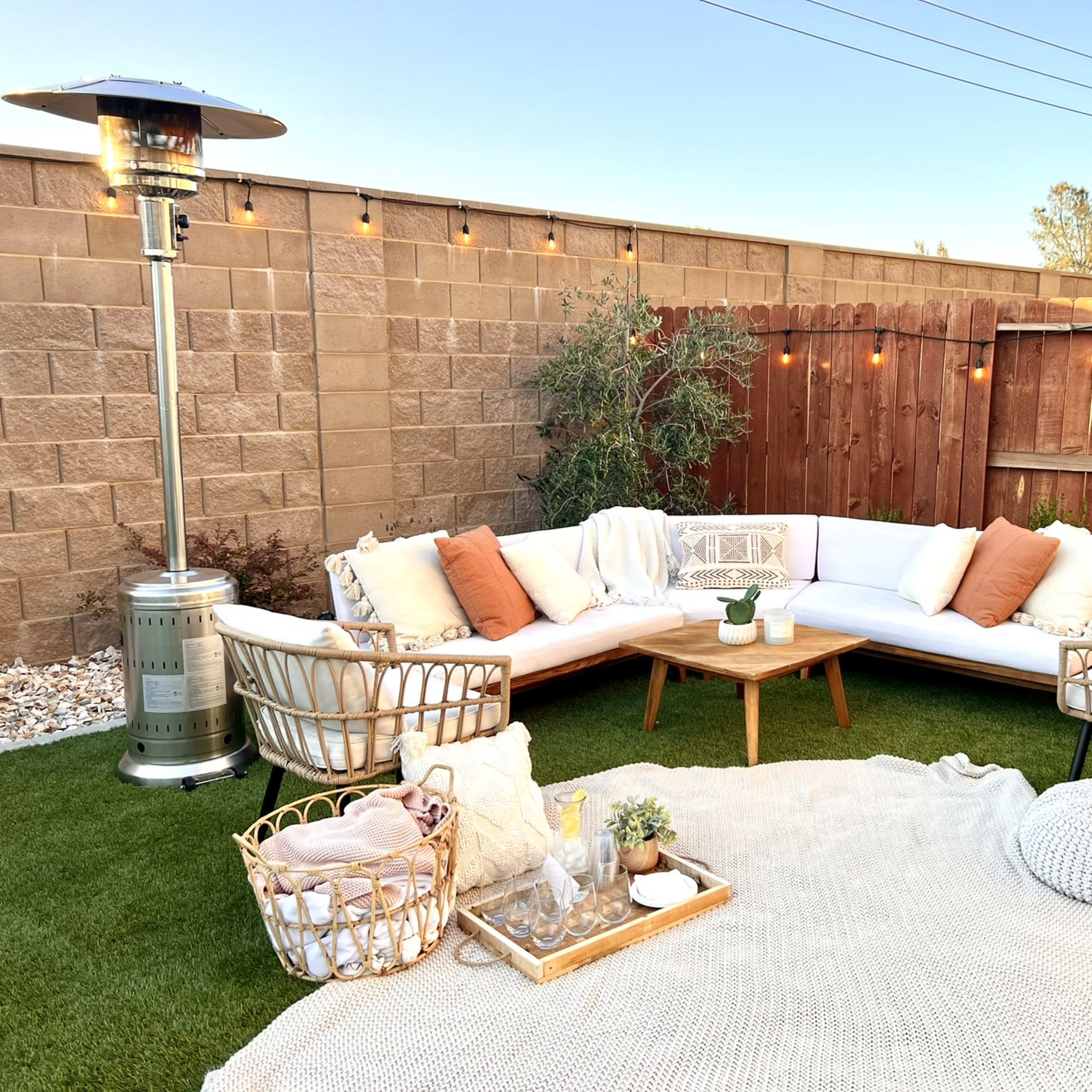 A cozy outdoor seating area features a sectional couch with throw pillows, a wooden coffee table, two chairs, and patio heaters, all surrounded by decorative lighting and greenery.