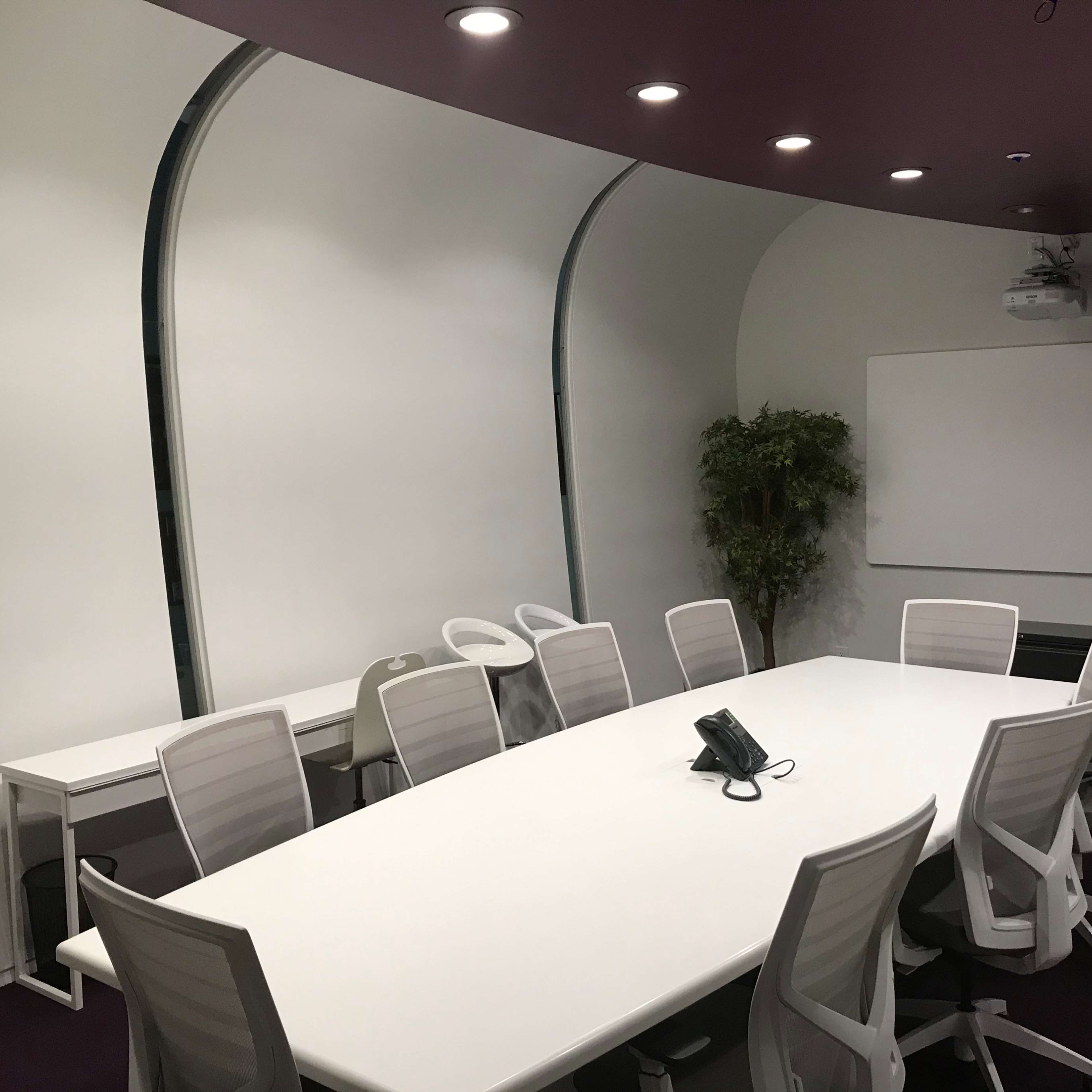 A modern conference room features a large white oval table surrounded by white chairs, with a projection screen and a potted plant in the corner.