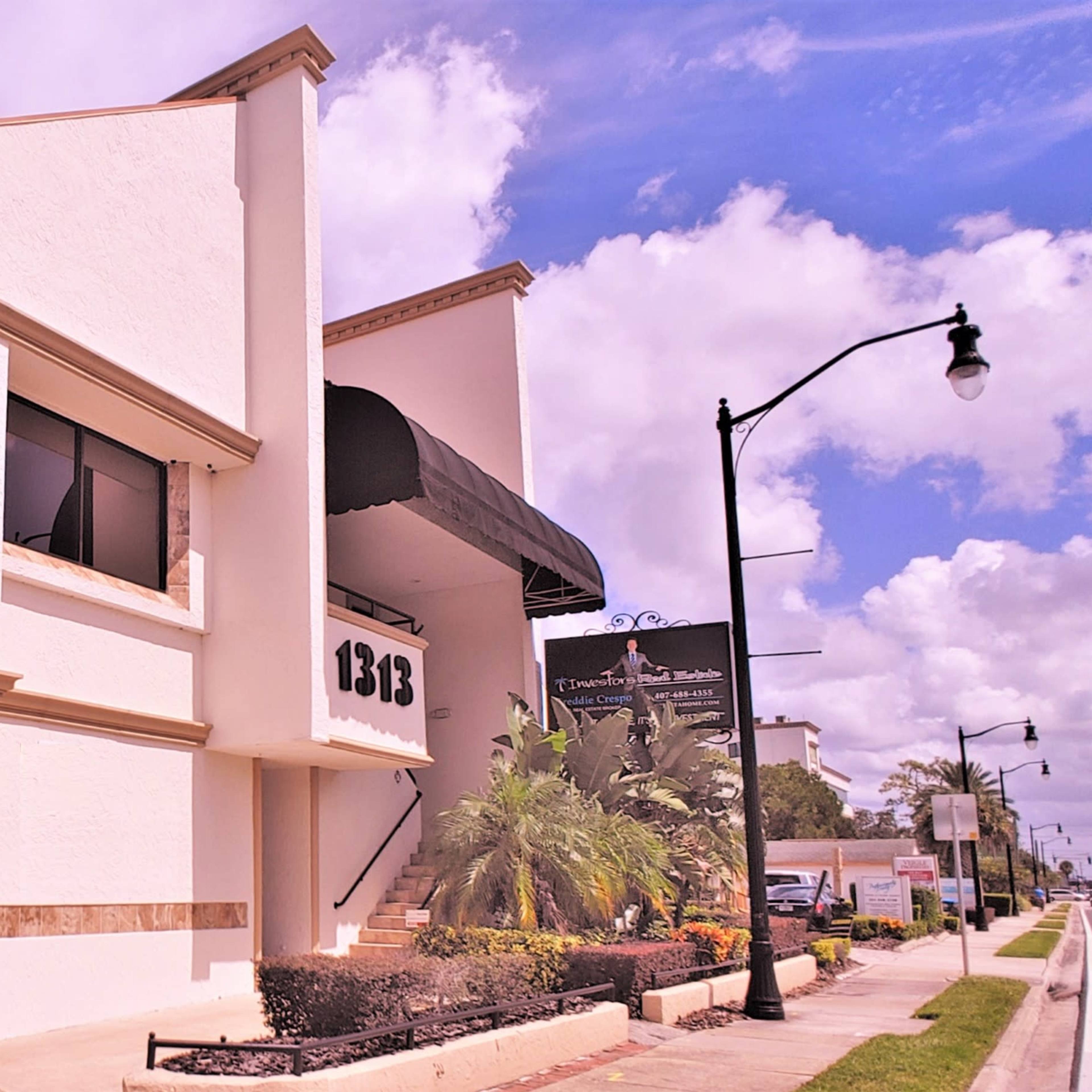 A two-story commercial building with a parking area underneath and palm trees, located on a sunny street.