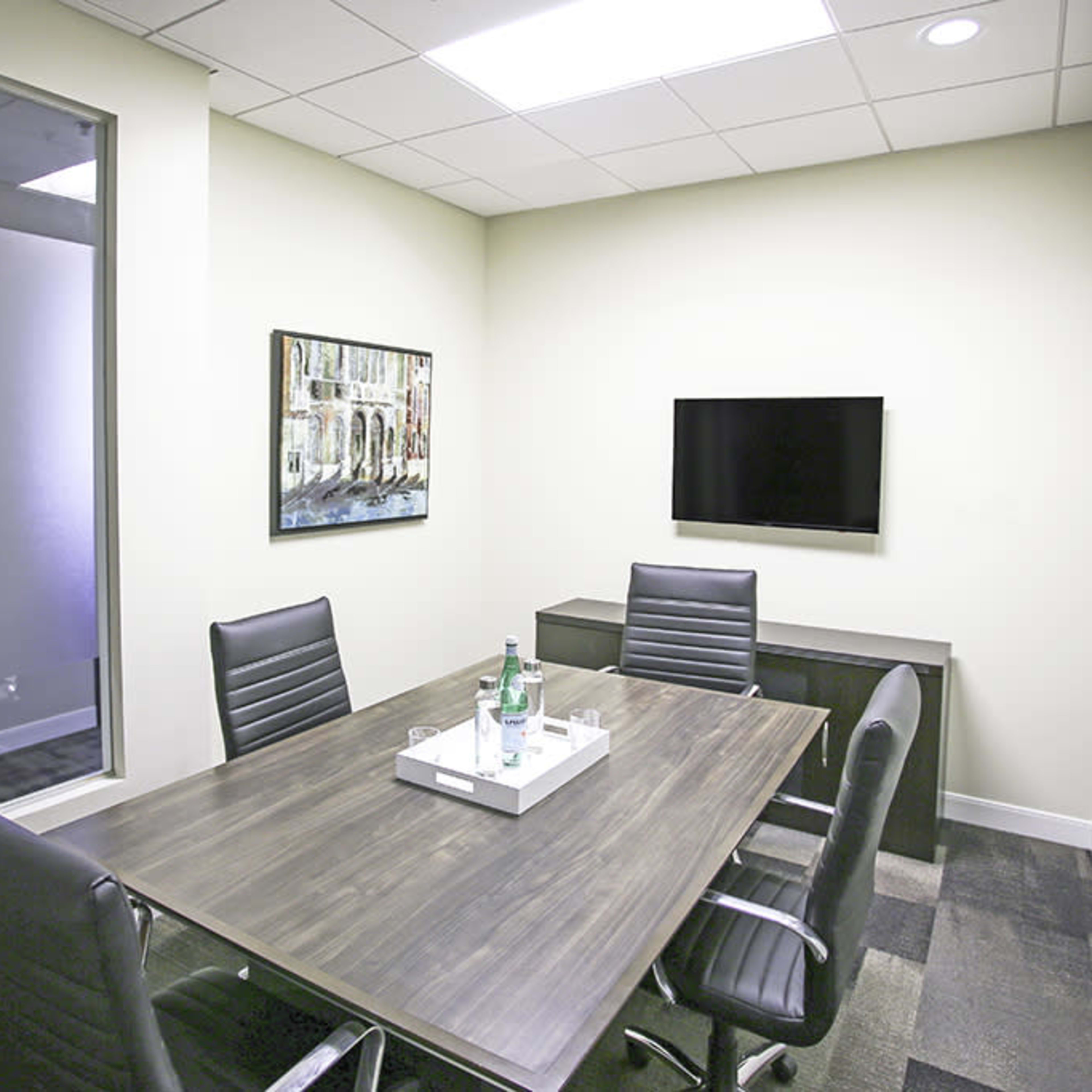 A small meeting room with a rectangular table, black leather chairs, a television mounted on the wall, and a minimalist decor.