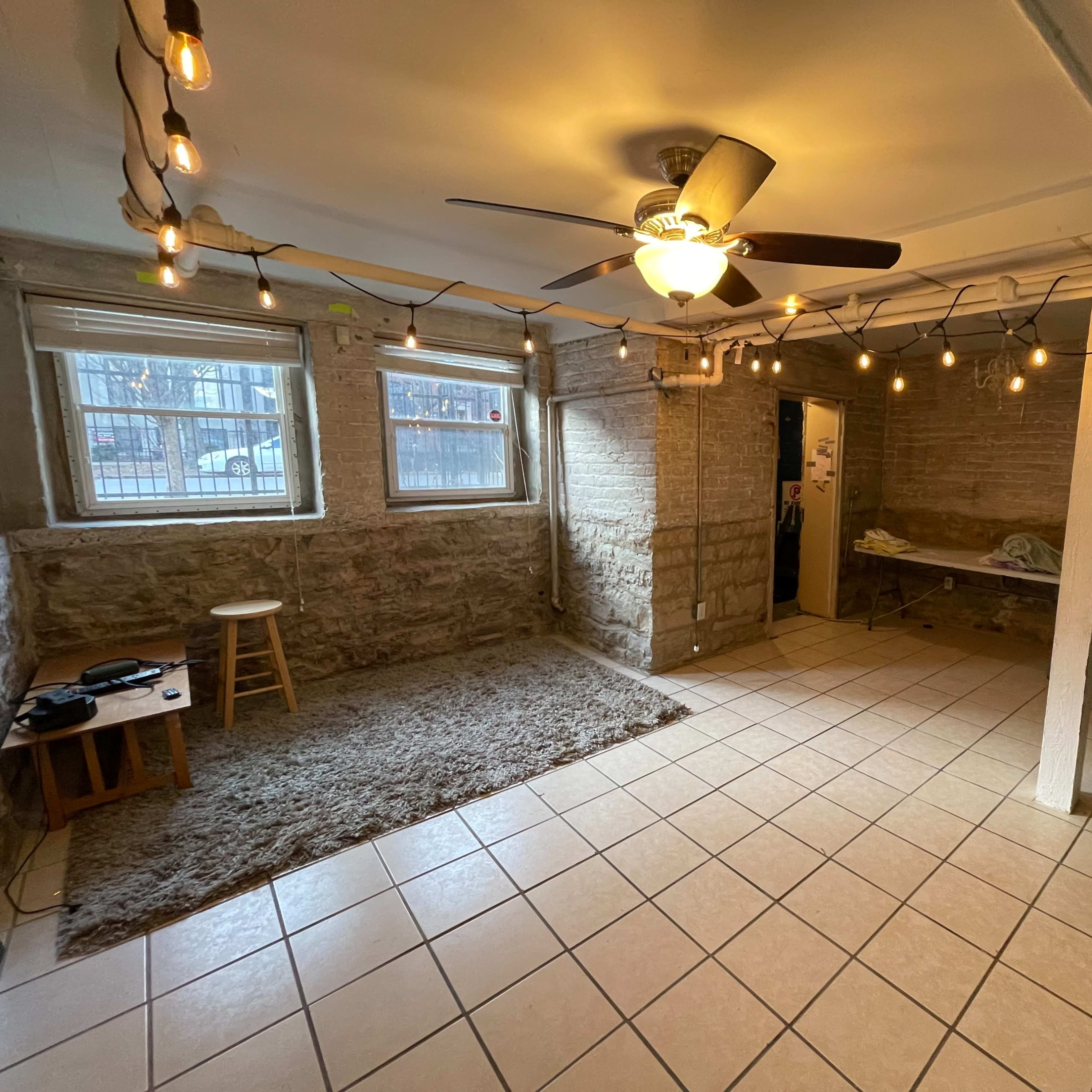 A basement room with a ceiling fan, string lights, tiled floor, and a few pieces of furniture including a stool and a table.