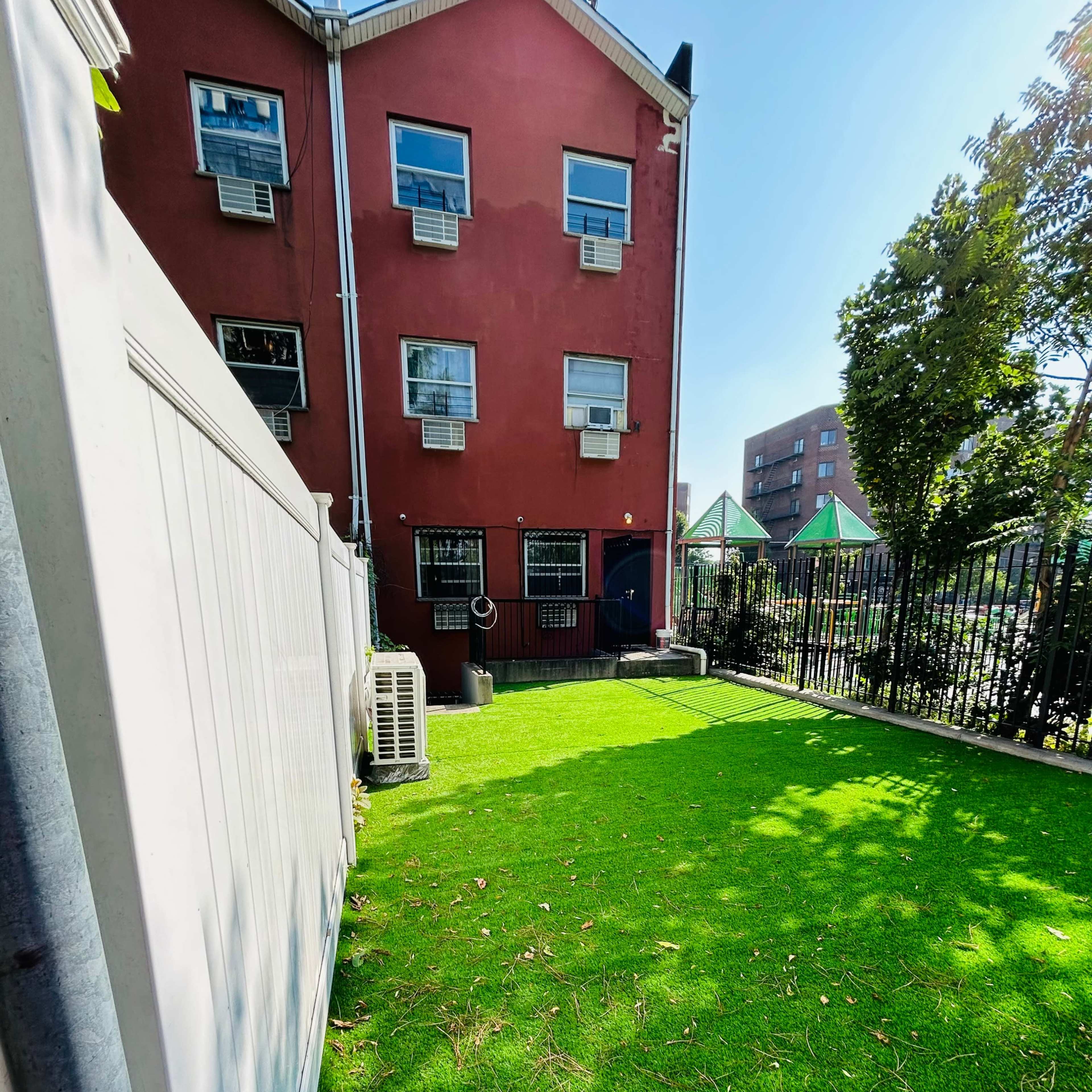 A small backyard features artificial grass and a red building with several air conditioning units.
