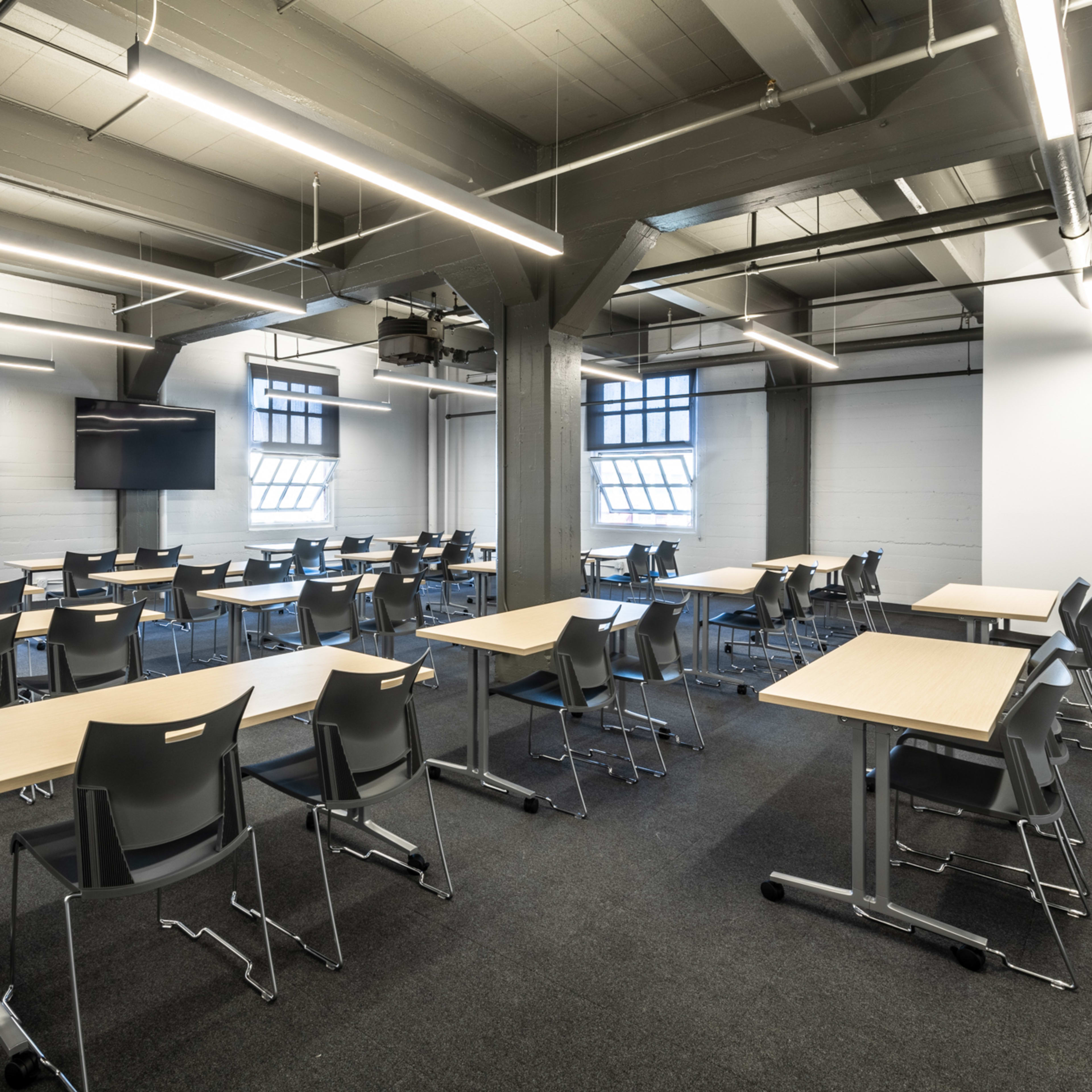 A modern classroom arranged with several tables and chairs facing a wall-mounted screen.