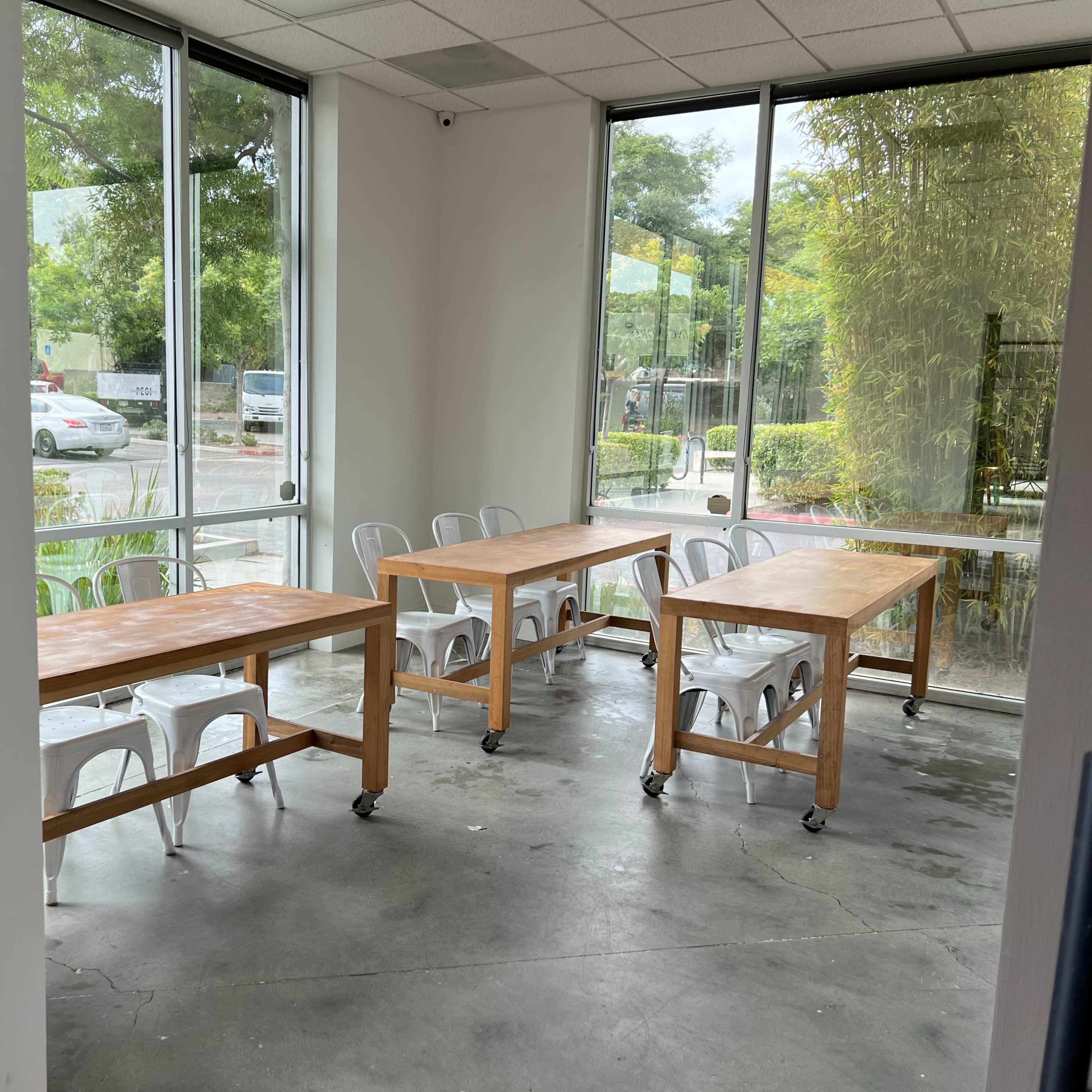 A room with four wooden tables on wheels and metal chairs, situated beside large windows that let in natural light.