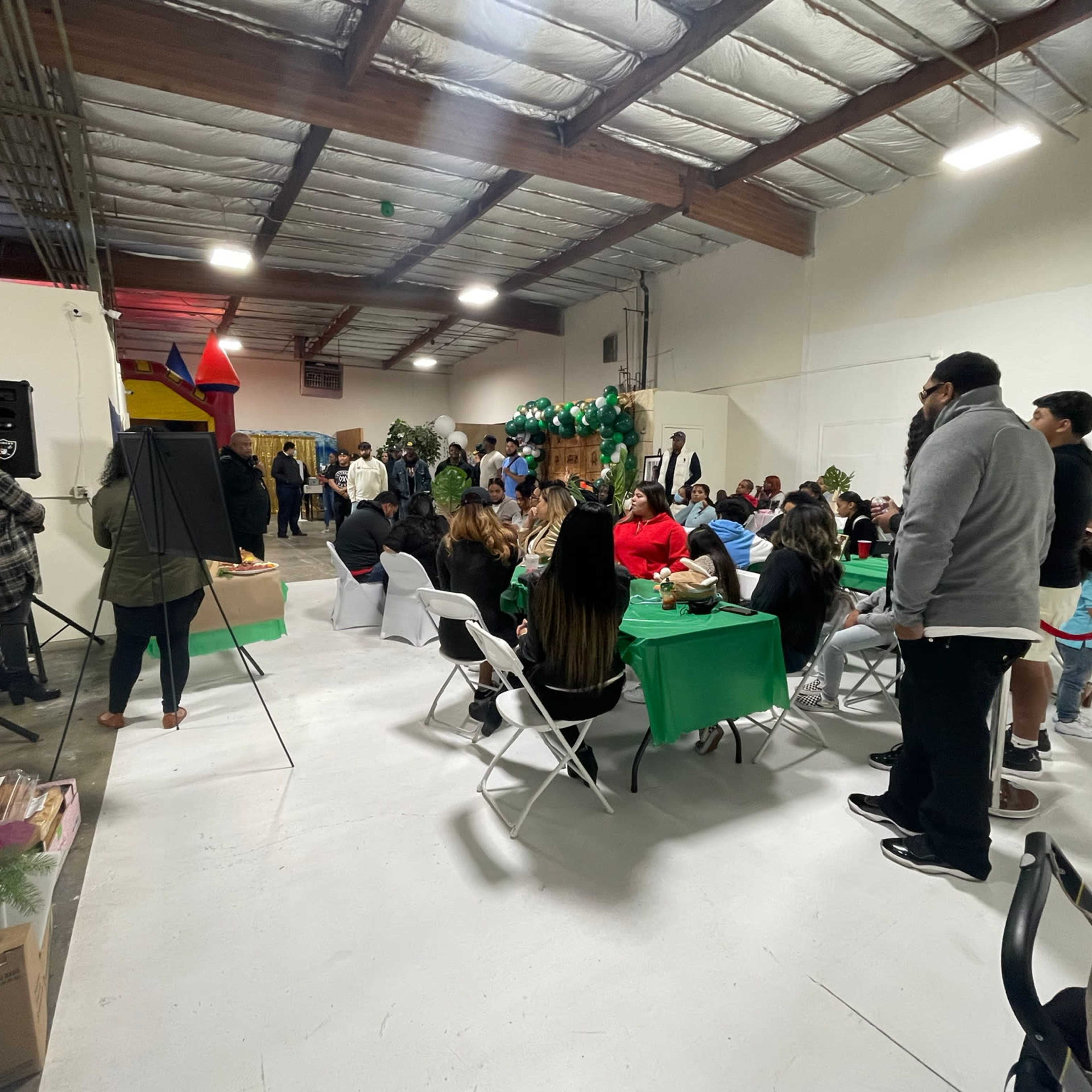 A crowd of people gathers around tables in a large indoor space decorated for an event, with some attendees interacting near a stage.