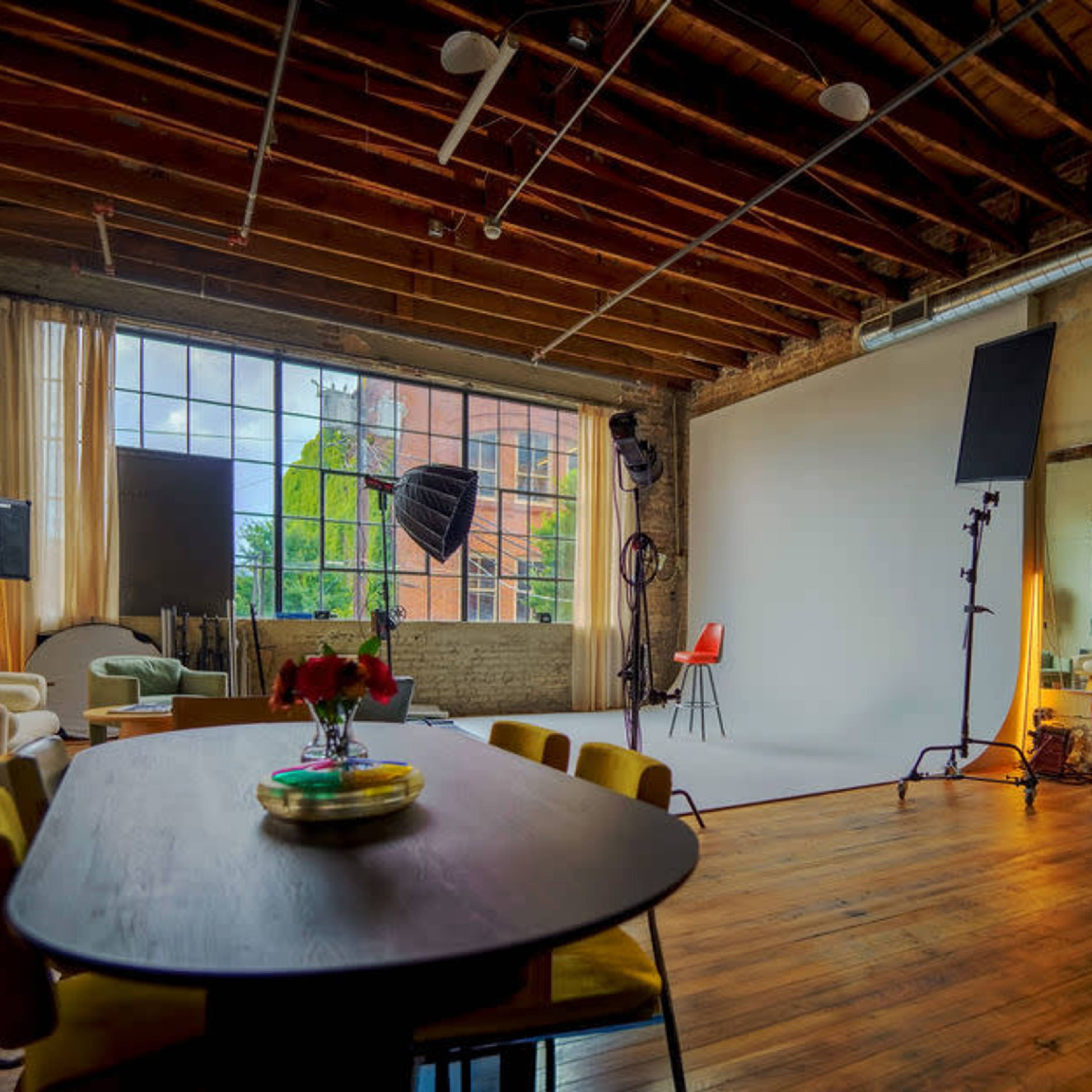 A spacious studio with wooden floors, large windows, and various furniture pieces, including a dining table and photography equipment.