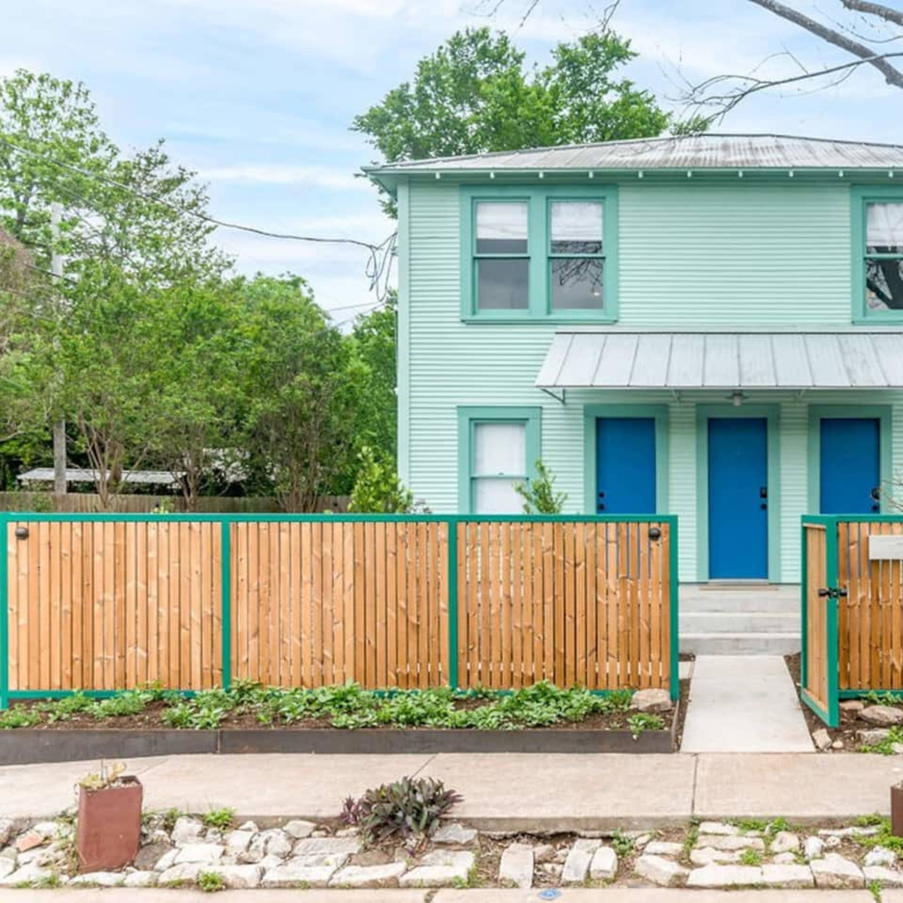 A two-story, mint green duplex with blue doors and a fenced front yard is shown.