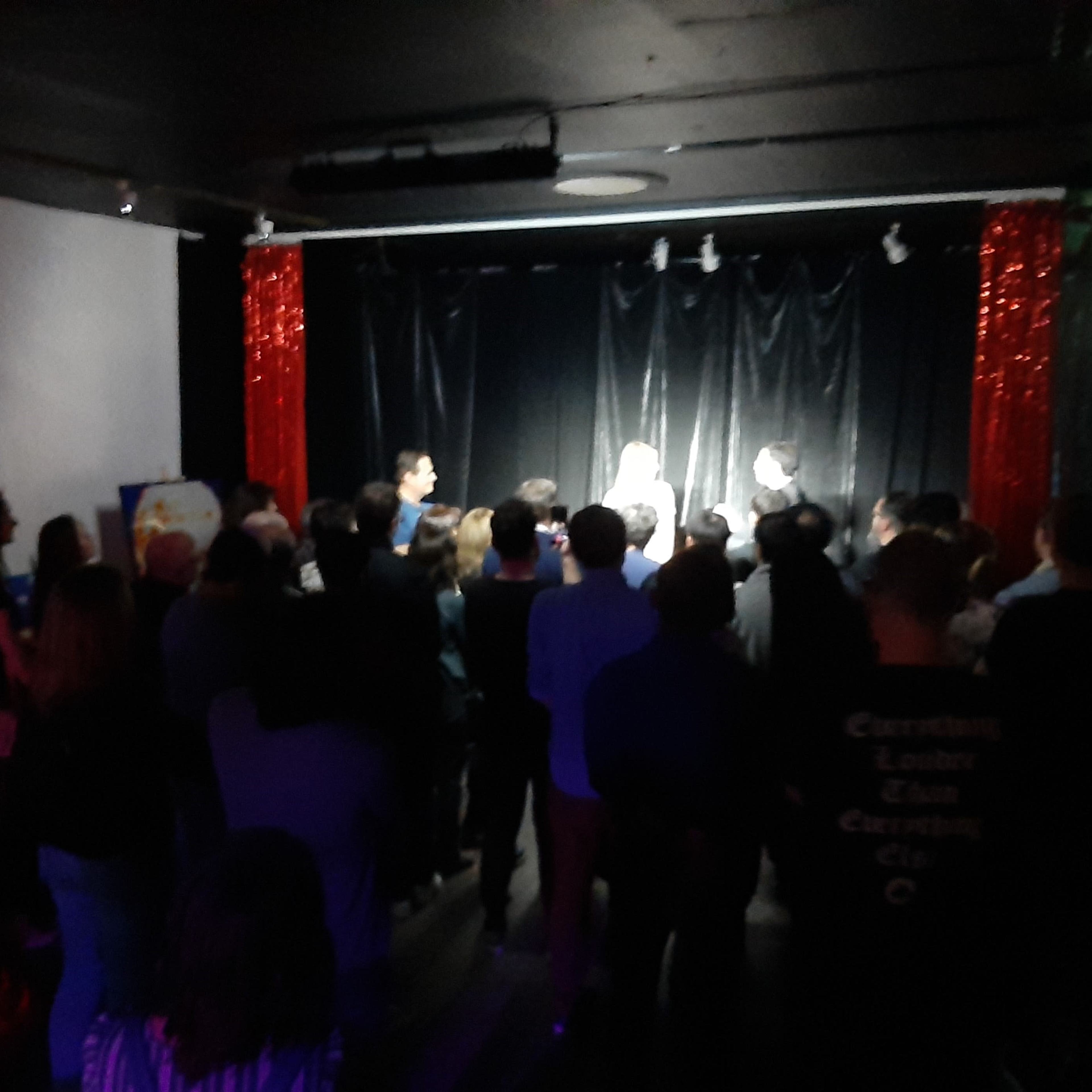 A crowd of people watches a performance on stage in a dimly lit venue adorned with red curtains.
