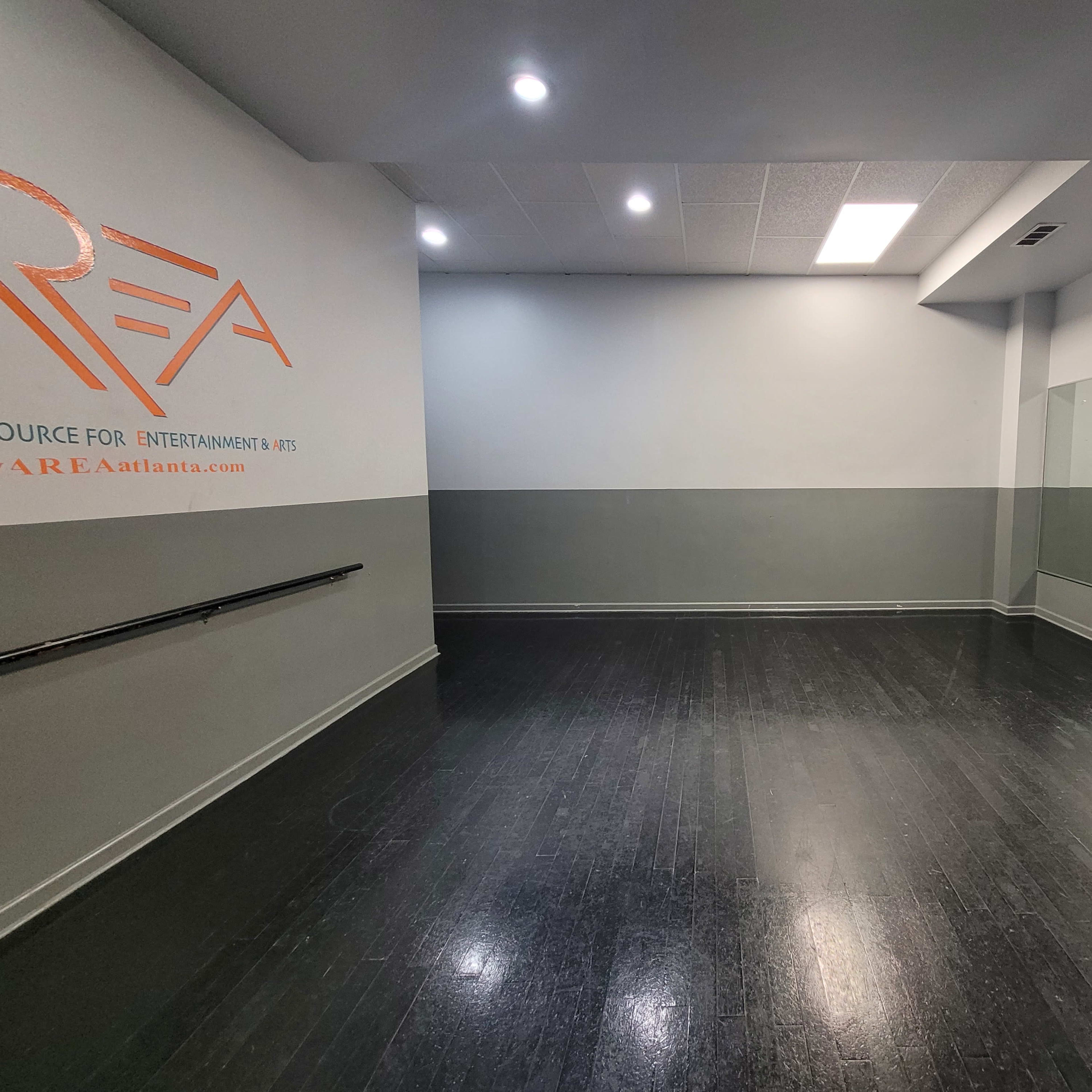 A dance studio with a mirrored wall, dark wooden flooring, and gray walls, illuminated by overhead lights.