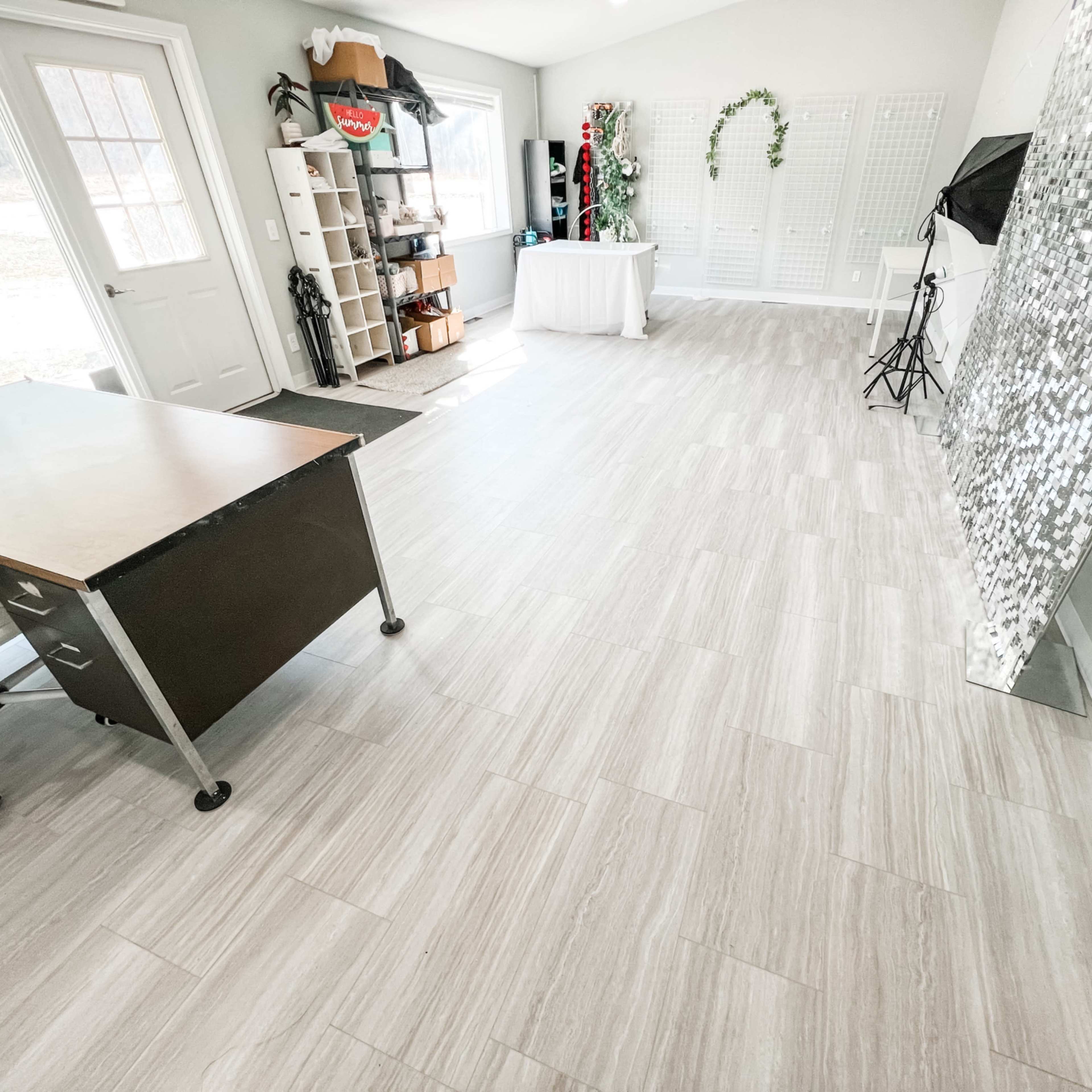 A spacious room with light-colored tiled flooring, a desk on wheels, and a mirrored wall, along with a white table and various props in the background.