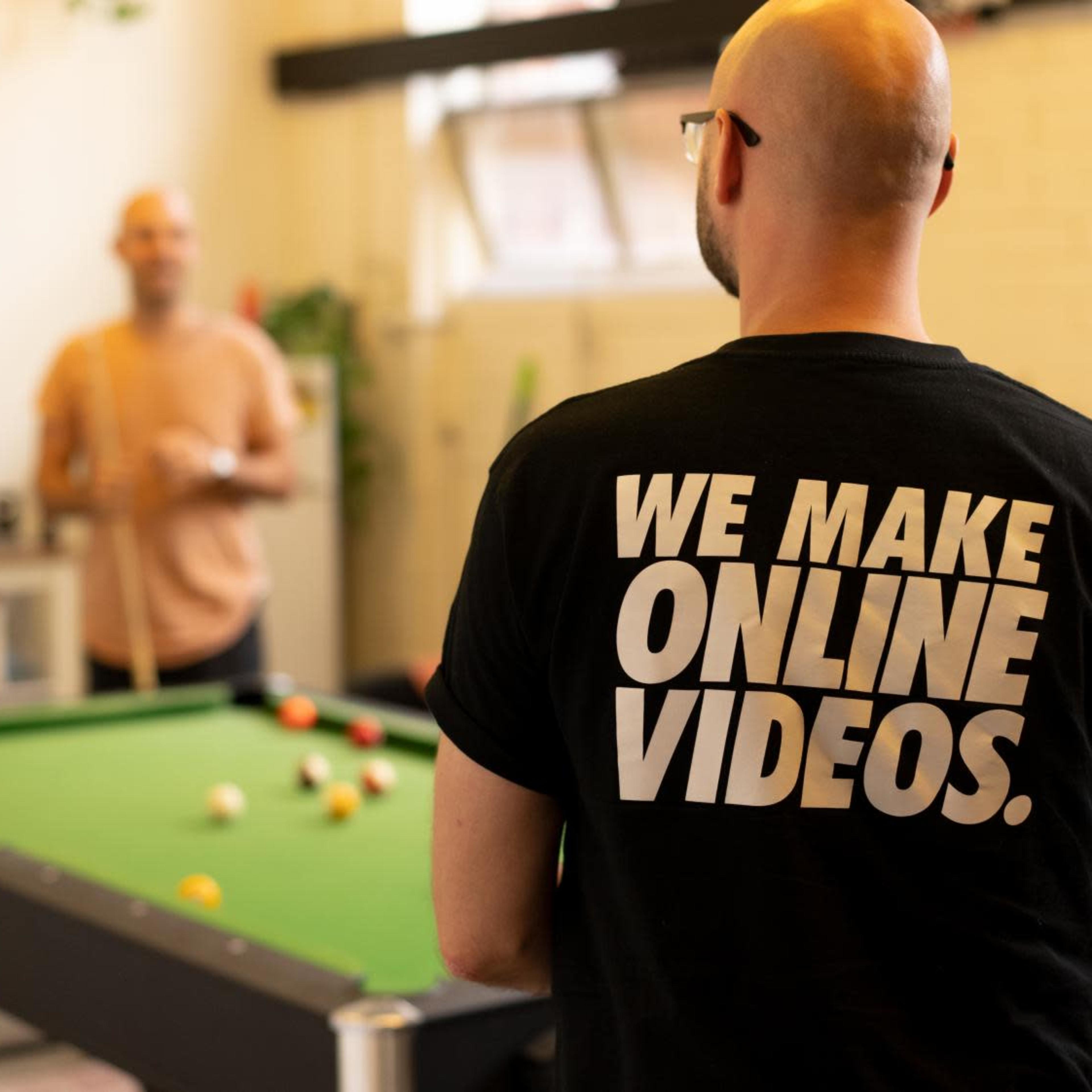Two men are in an indoor space, one standing at a pool table and the other facing away, wearing a shirt that says "WE MAKE ONLINE VIDEOS."