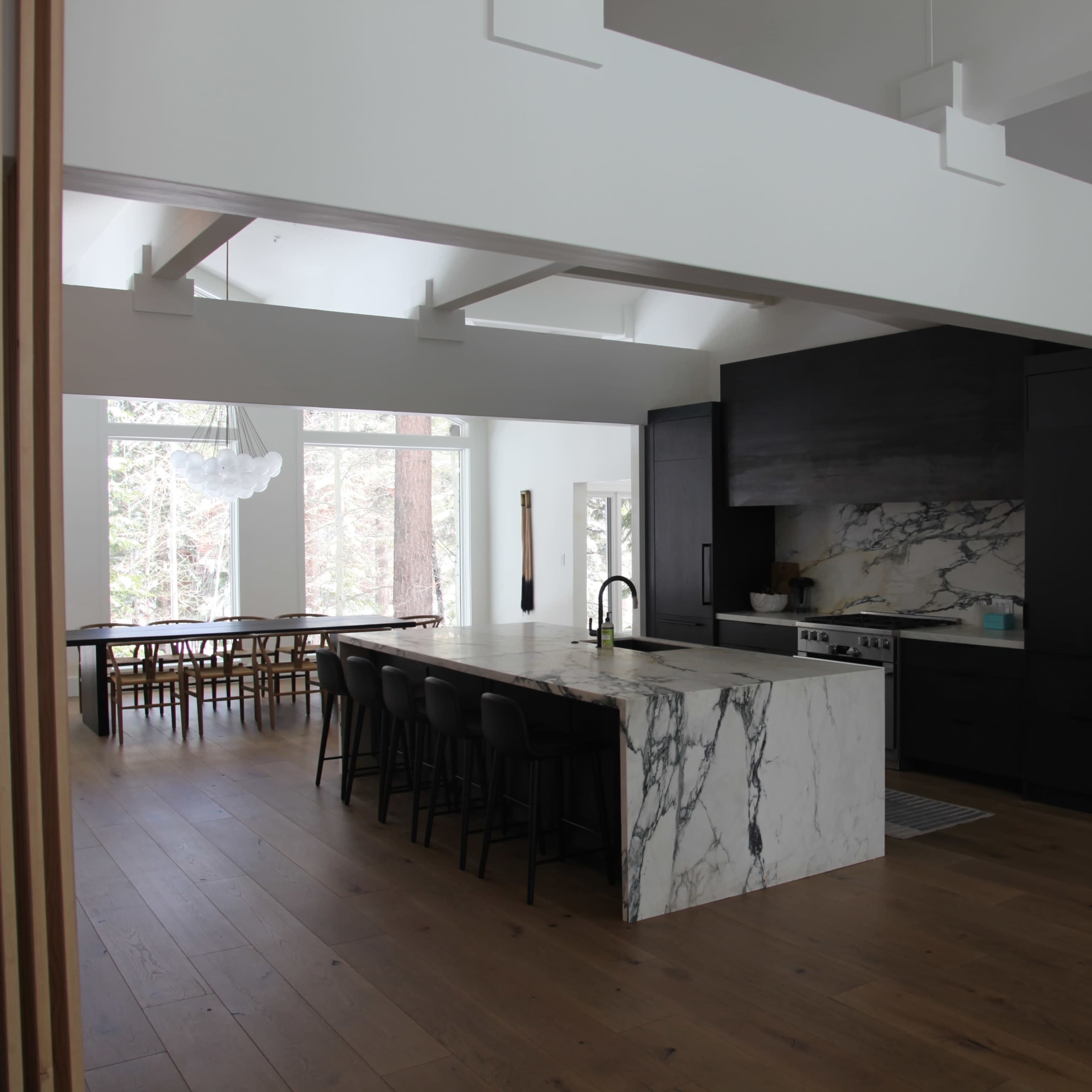 A modern kitchen features a large marble island, black cabinetry, and a dining area with a long table against large windows.