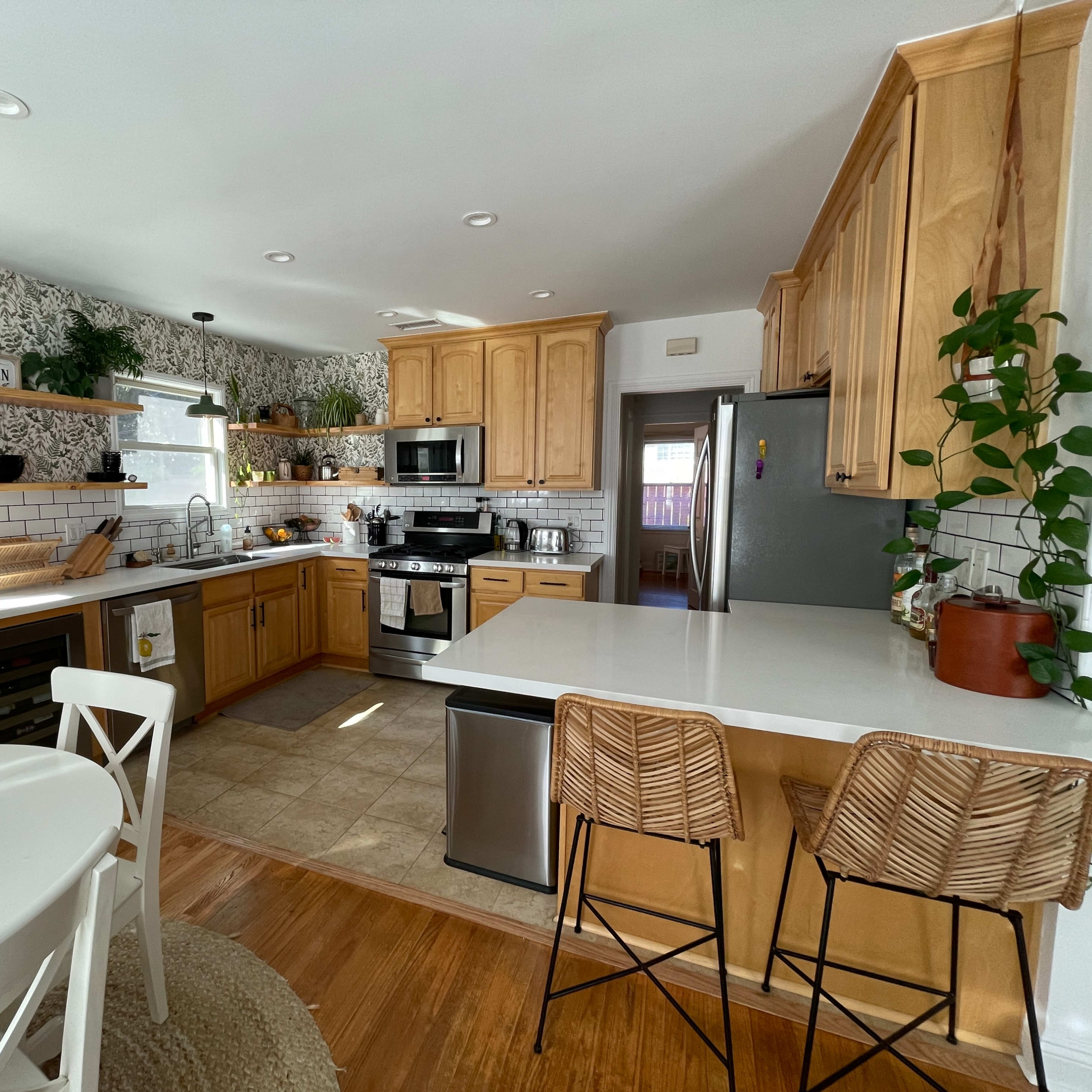 A modern kitchen with wooden cabinets, stainless steel appliances, a white countertop, and a dining area with a round table and bar stools.