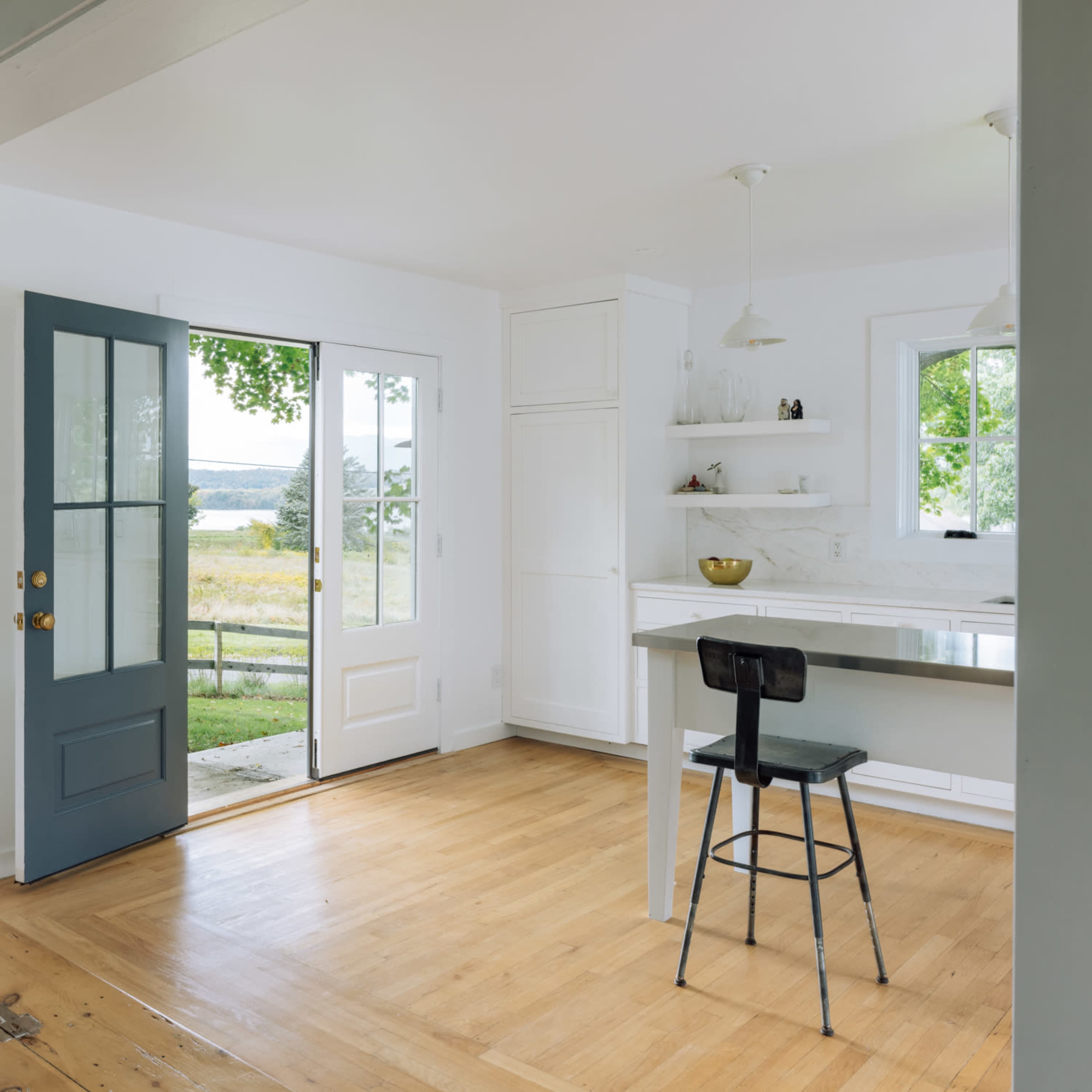 A bright, minimalist kitchen opens to an outdoor view through a large door, featuring wooden floors and simple furnishings.