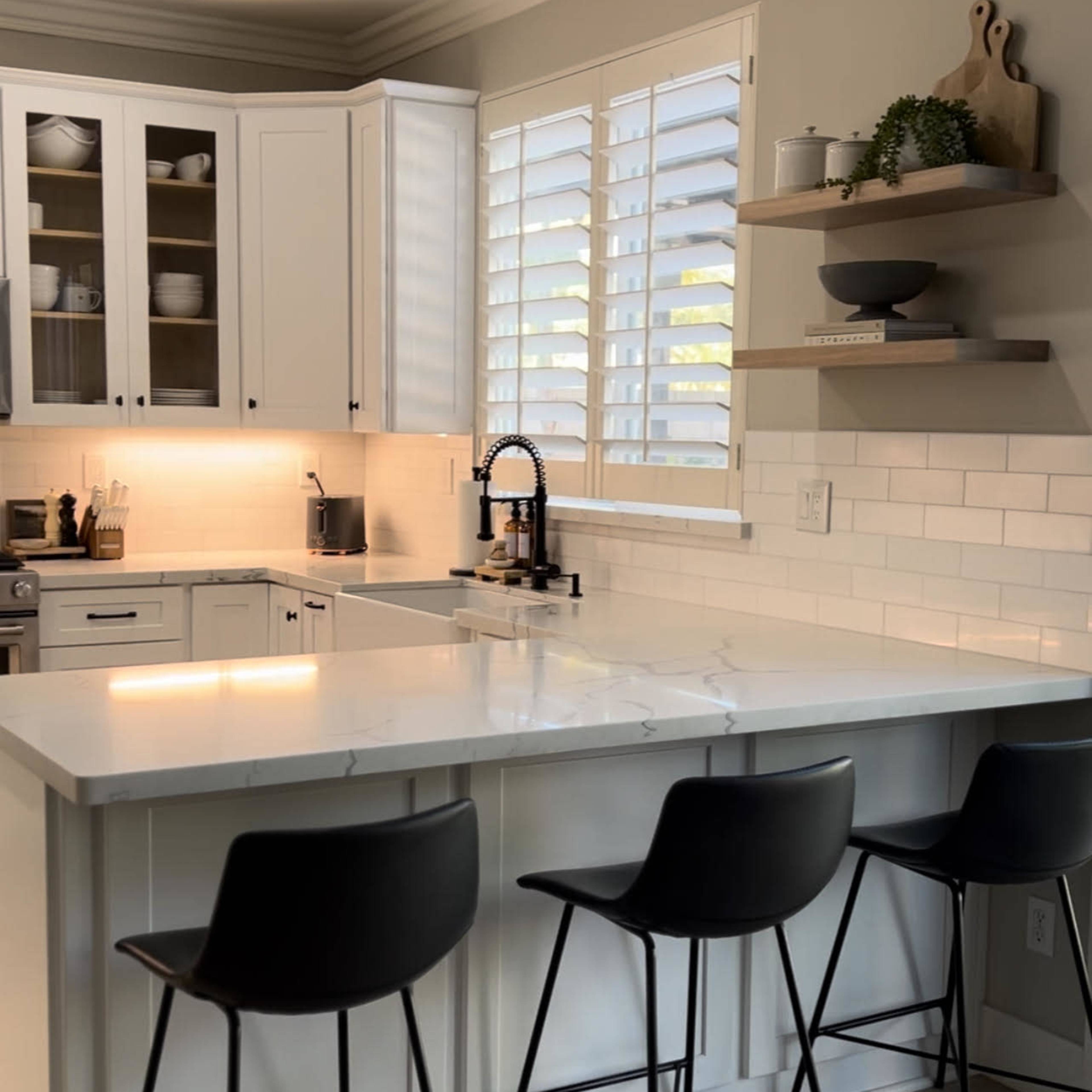 A modern kitchen features white cabinetry, a central island with marble countertops, and three black bar stools.