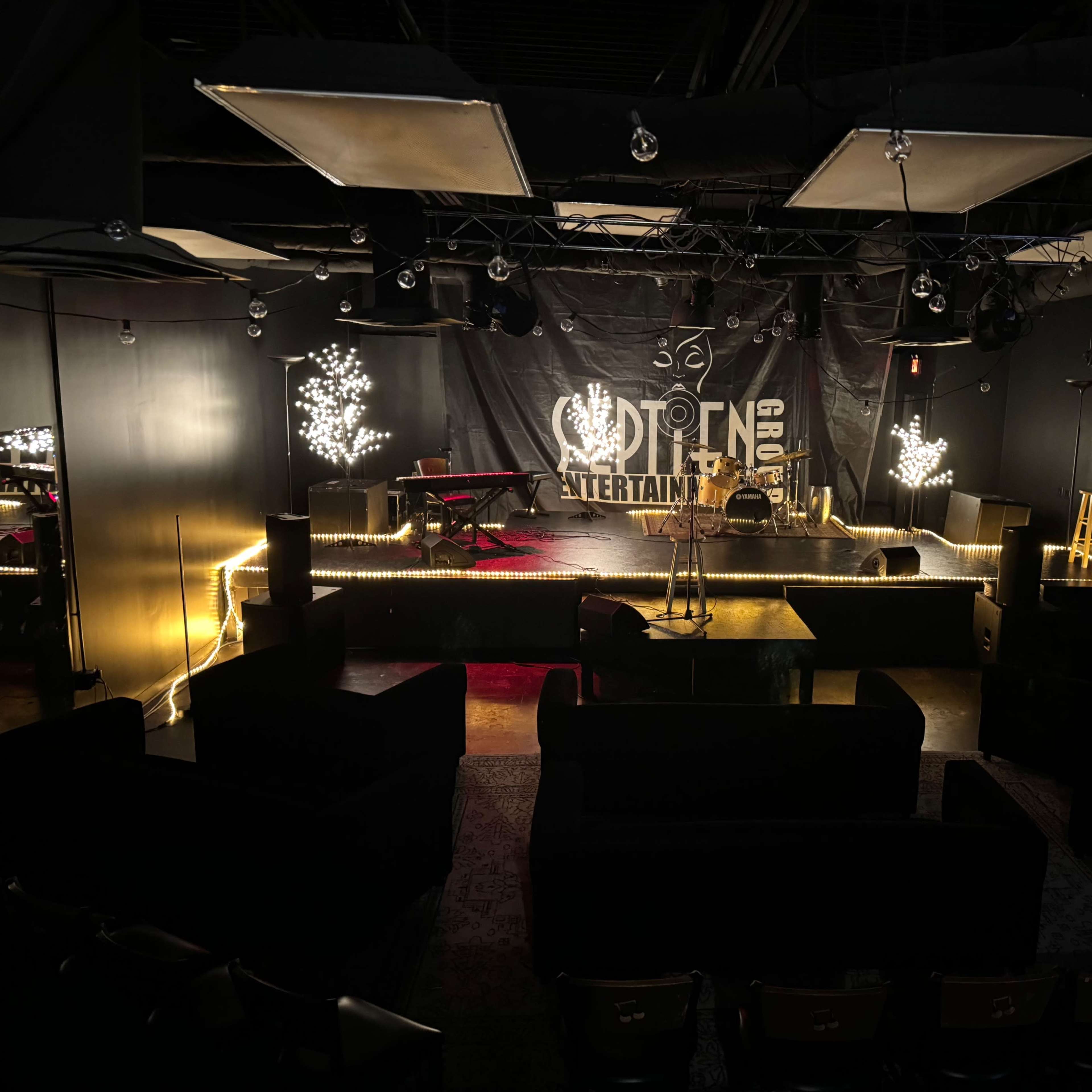 A stage area in a dark venue, featuring a microphone stand, a drum set, and seating arrangements with couches and chairs.