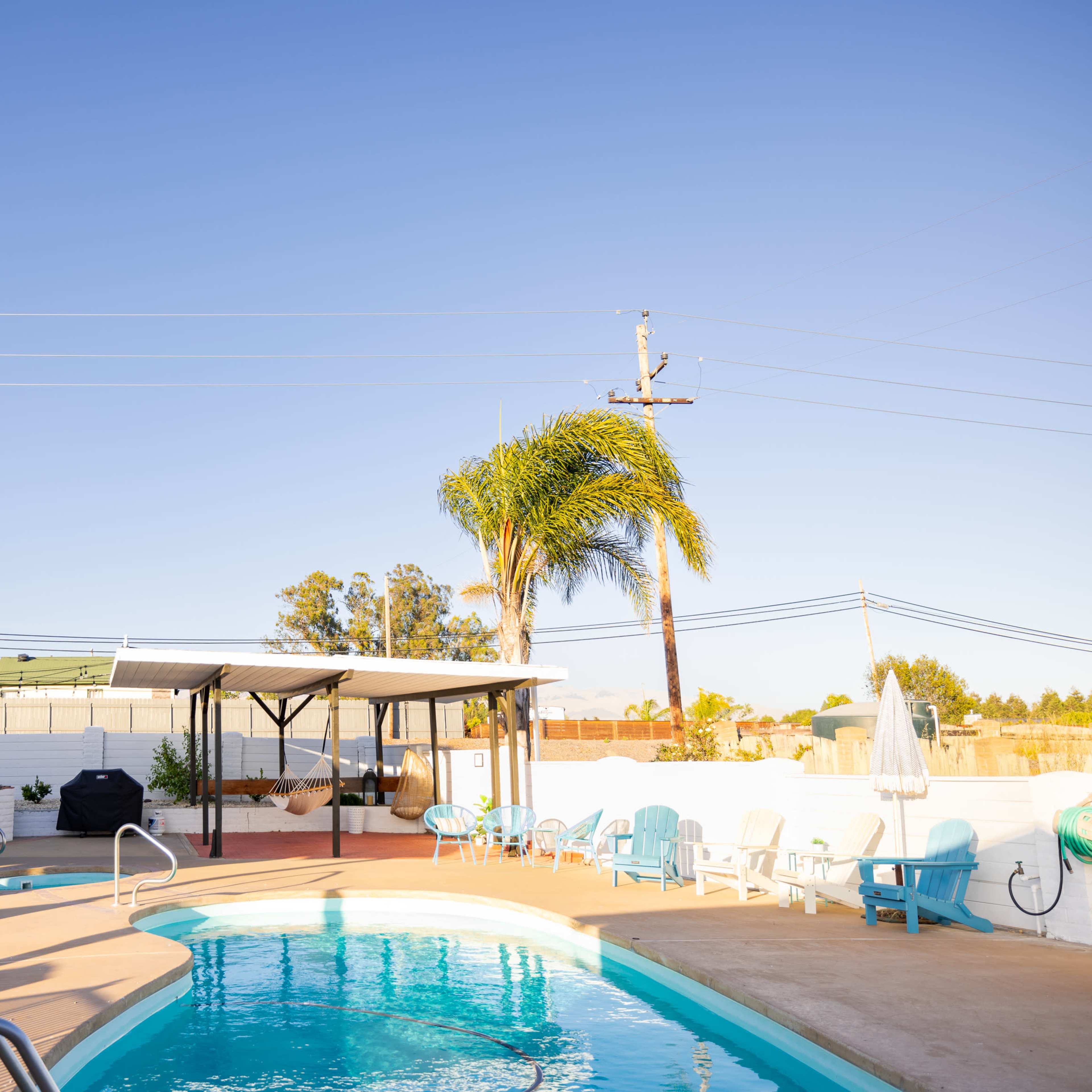A swimming pool is surrounded by patio furniture and shaded areas, with a clear blue sky and palm trees in the background.