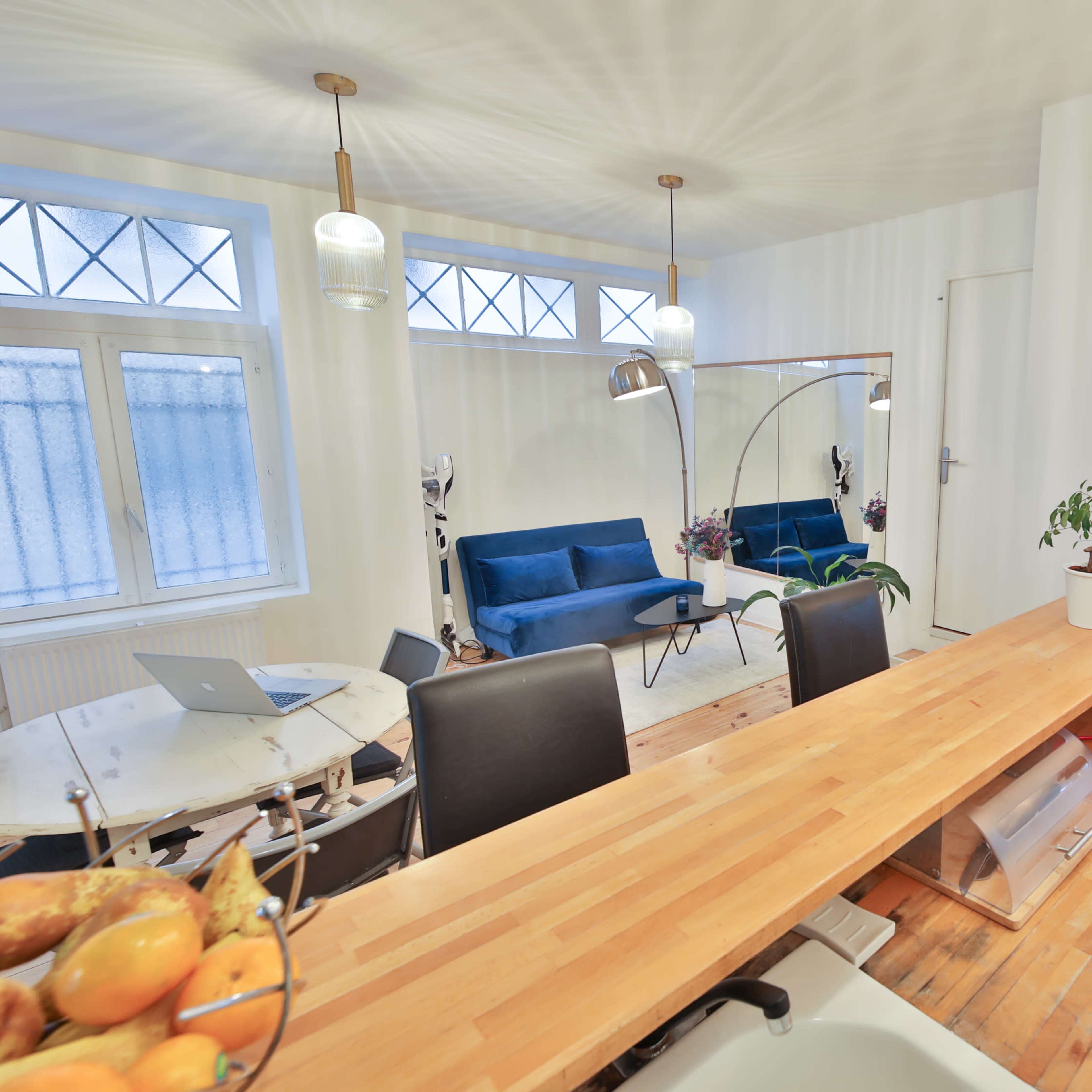 A modern kitchen and dining area featuring a wooden countertop, blue upholstered sofas, a round dining table, and large windows.