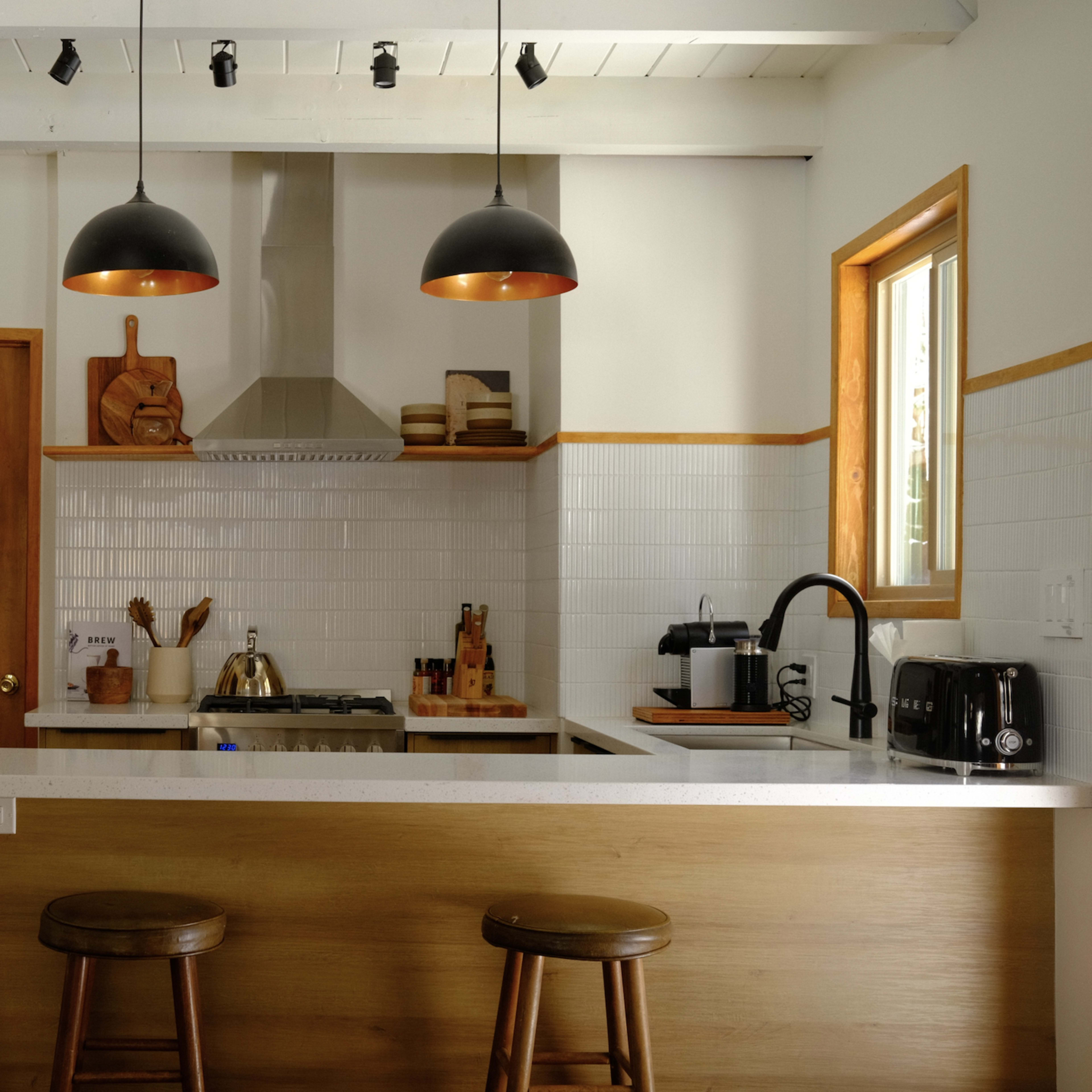 A modern kitchen with a wooden bar, two stools, stainless steel appliances, and black pendant lights.