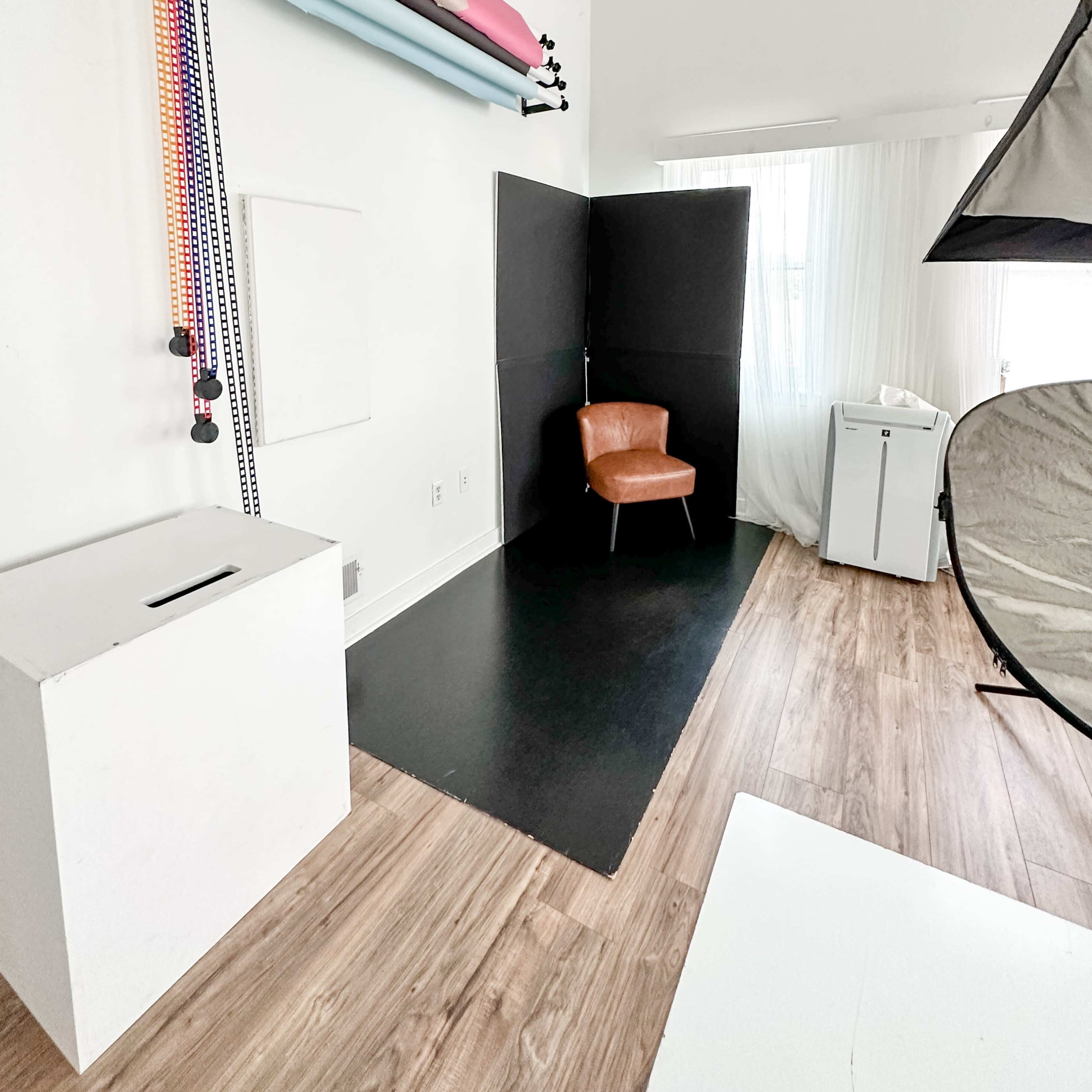 A photography studio with a brown chair positioned next to a backdrop stand, rollers of colored paper, and a softbox.