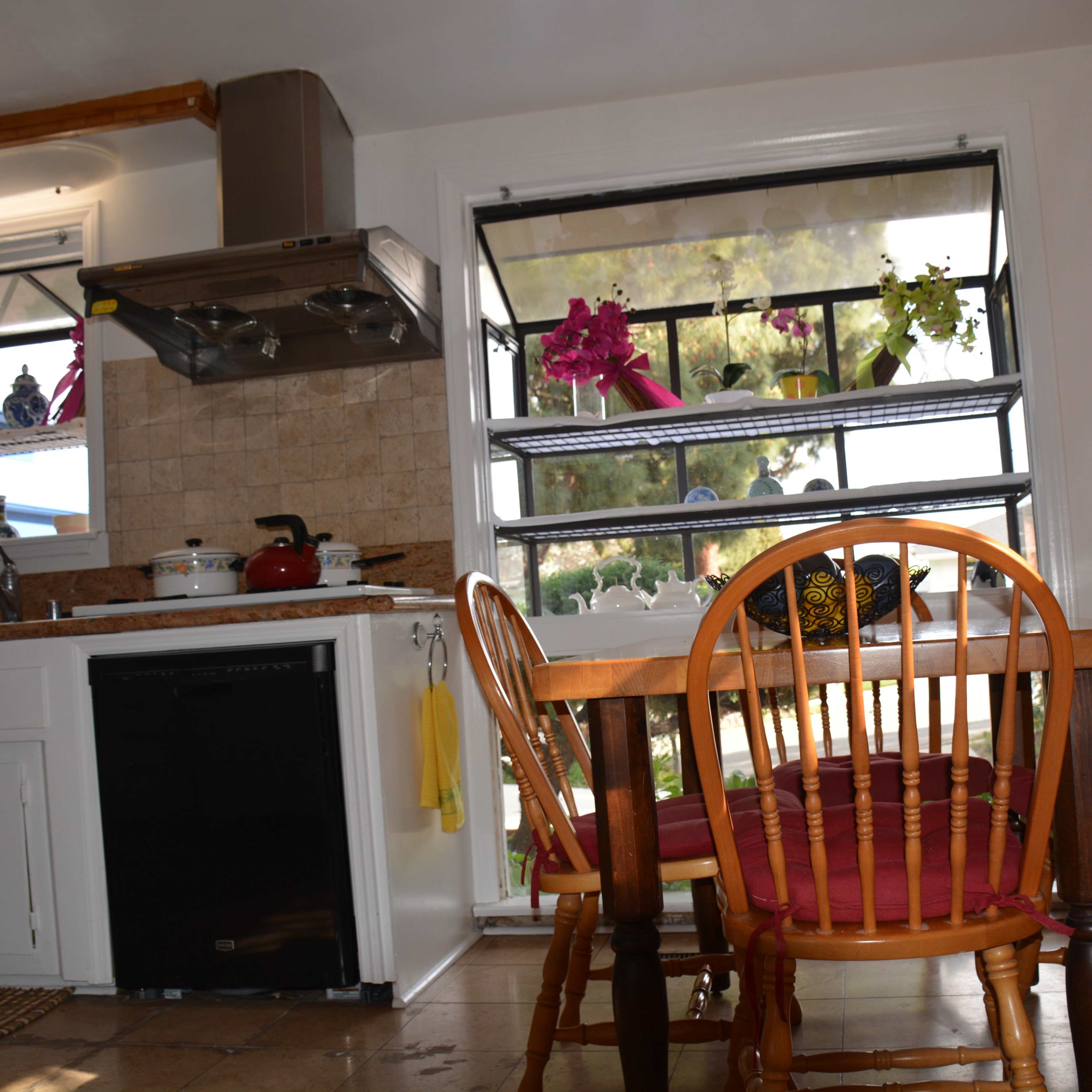 A kitchen with a wooden dining table and chairs, a stove and sink area, and large windows displaying potted plants.