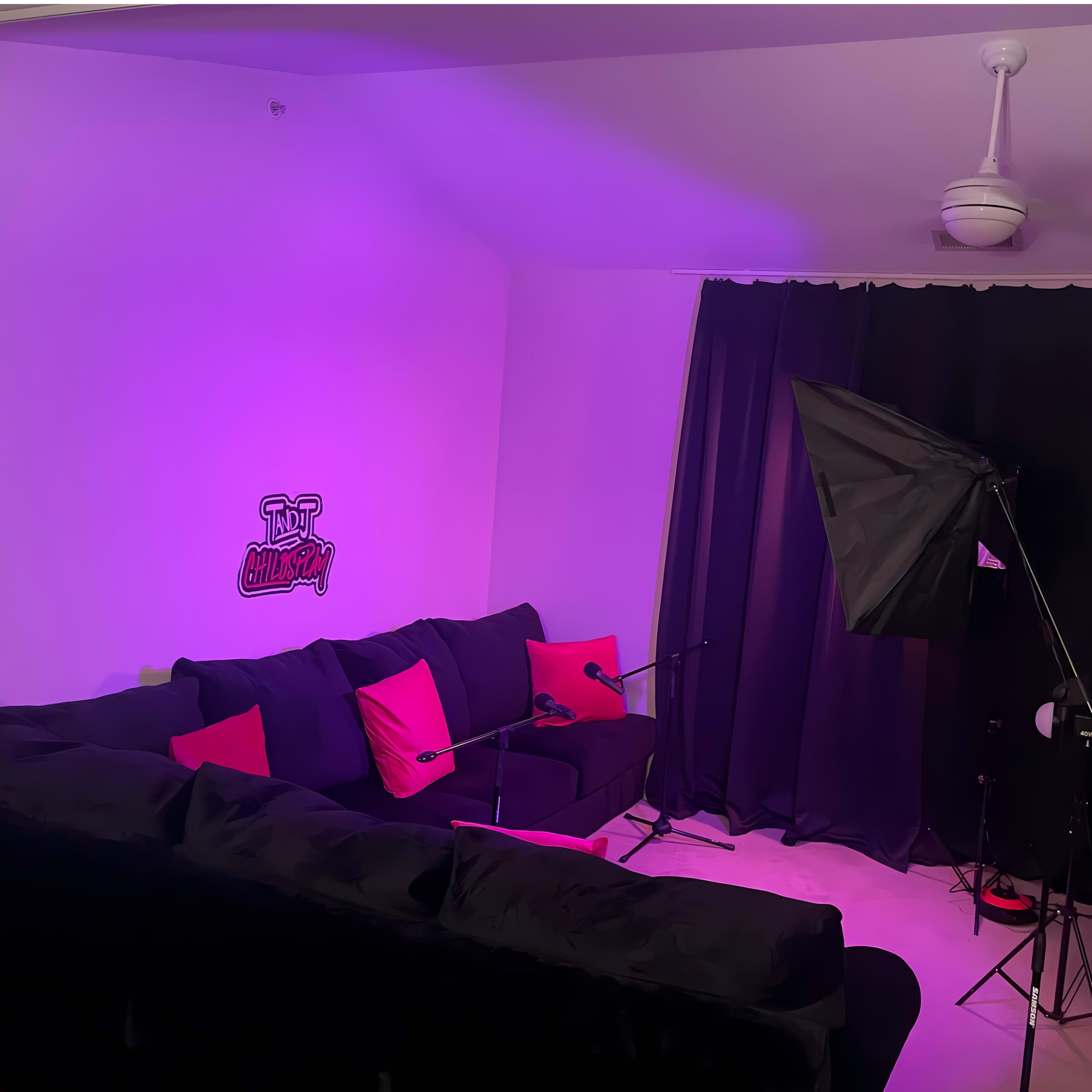 A living room features a large black couch adorned with red cushions, against a backdrop of purple lighting and black curtains, with a microphone setup and a camera on a stand.