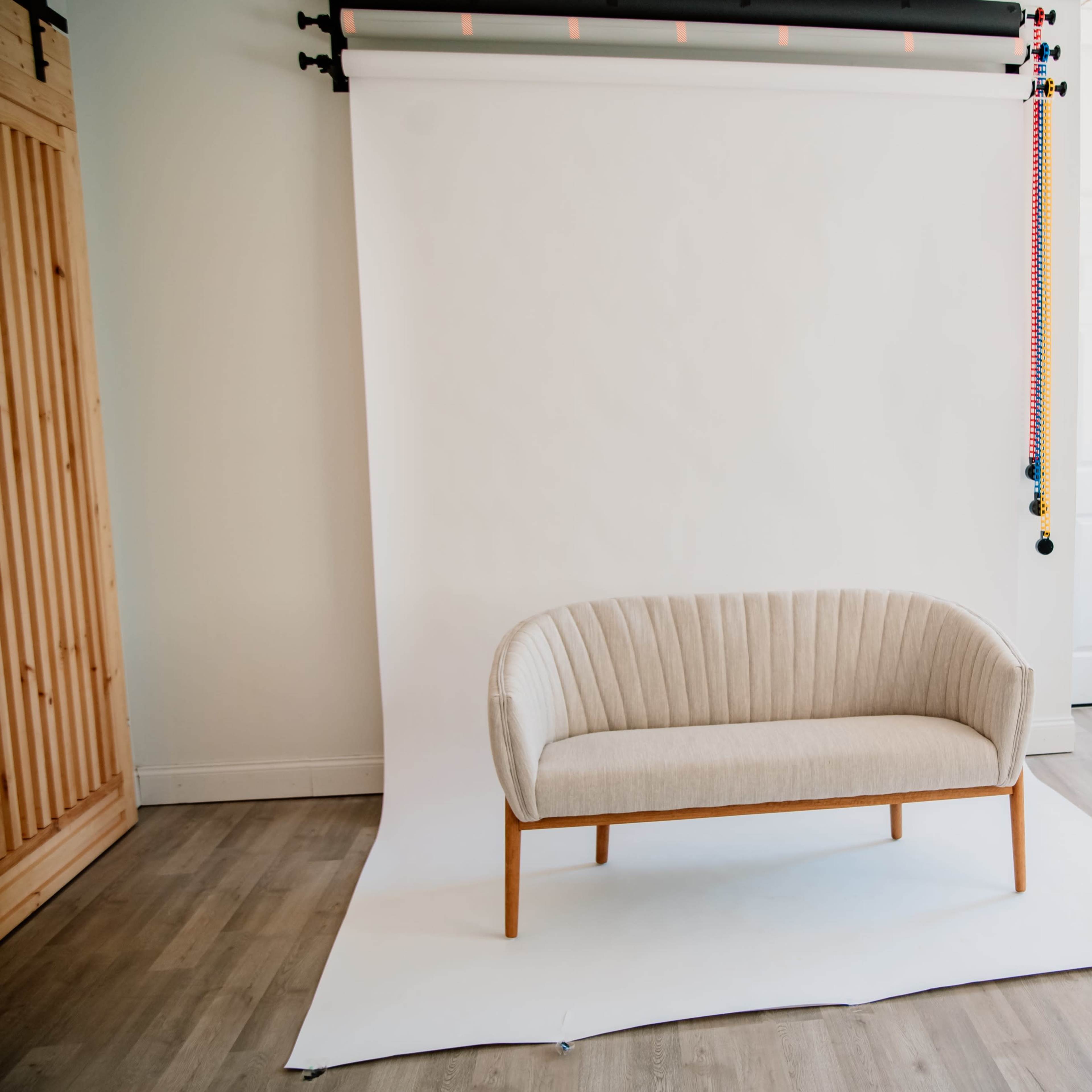 A beige upholstered couch with wood legs is centered in a bright, minimalistic room with white walls and hardwood flooring.