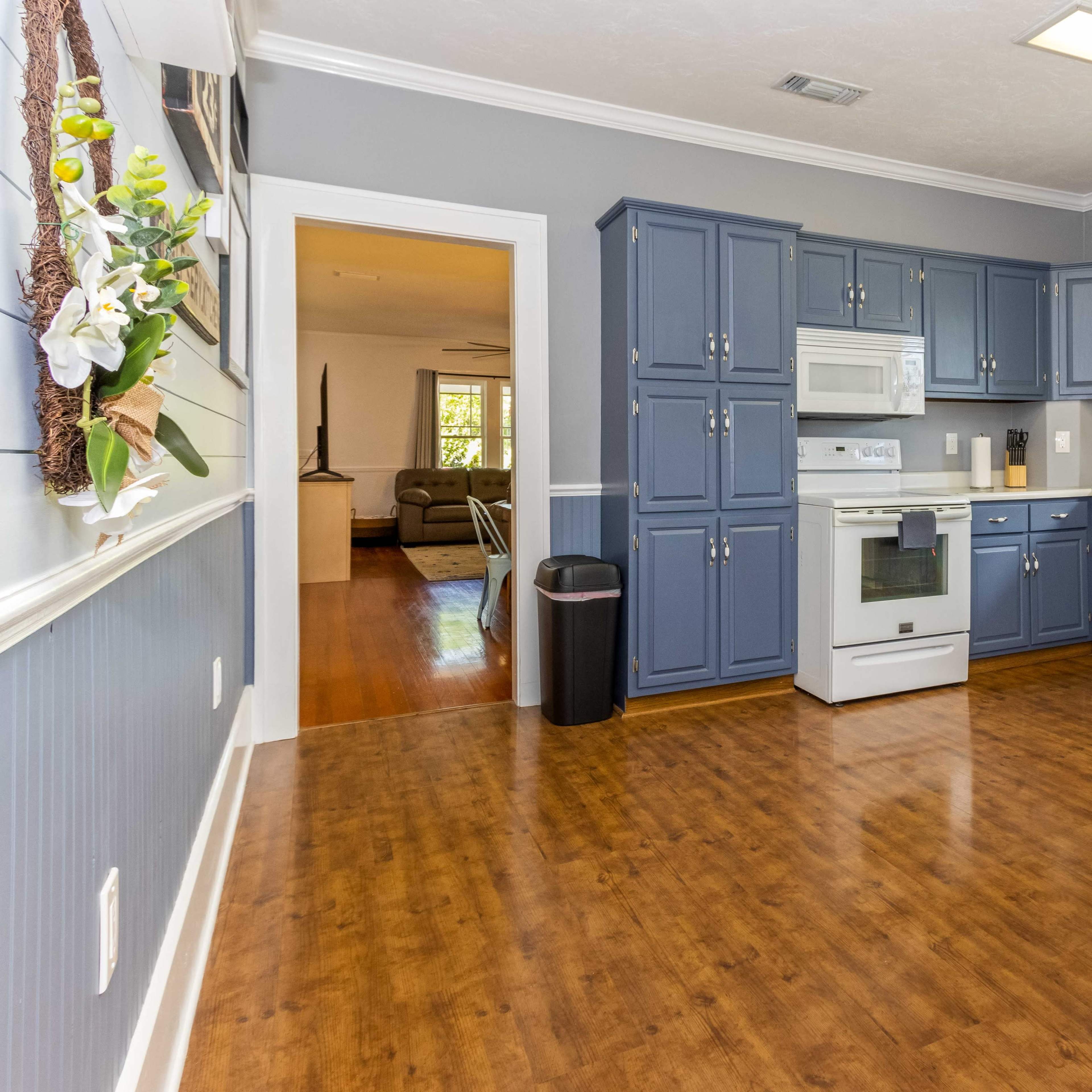A kitchen with blue cabinets, a white microwave and oven, and a view of a living room through an open doorway.