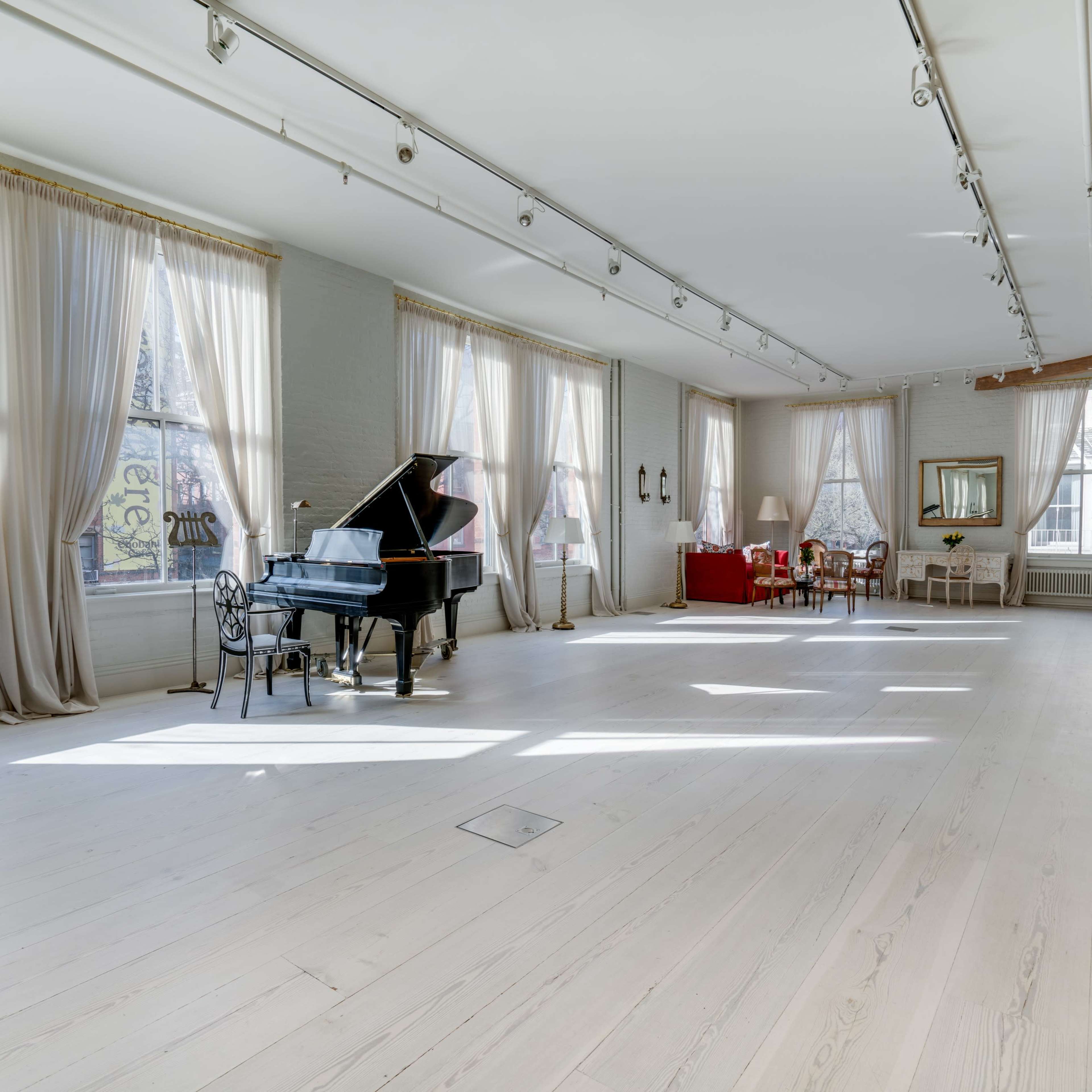 A spacious room features a grand piano, multiple large windows with drapes, and a seating area with elegant furniture.