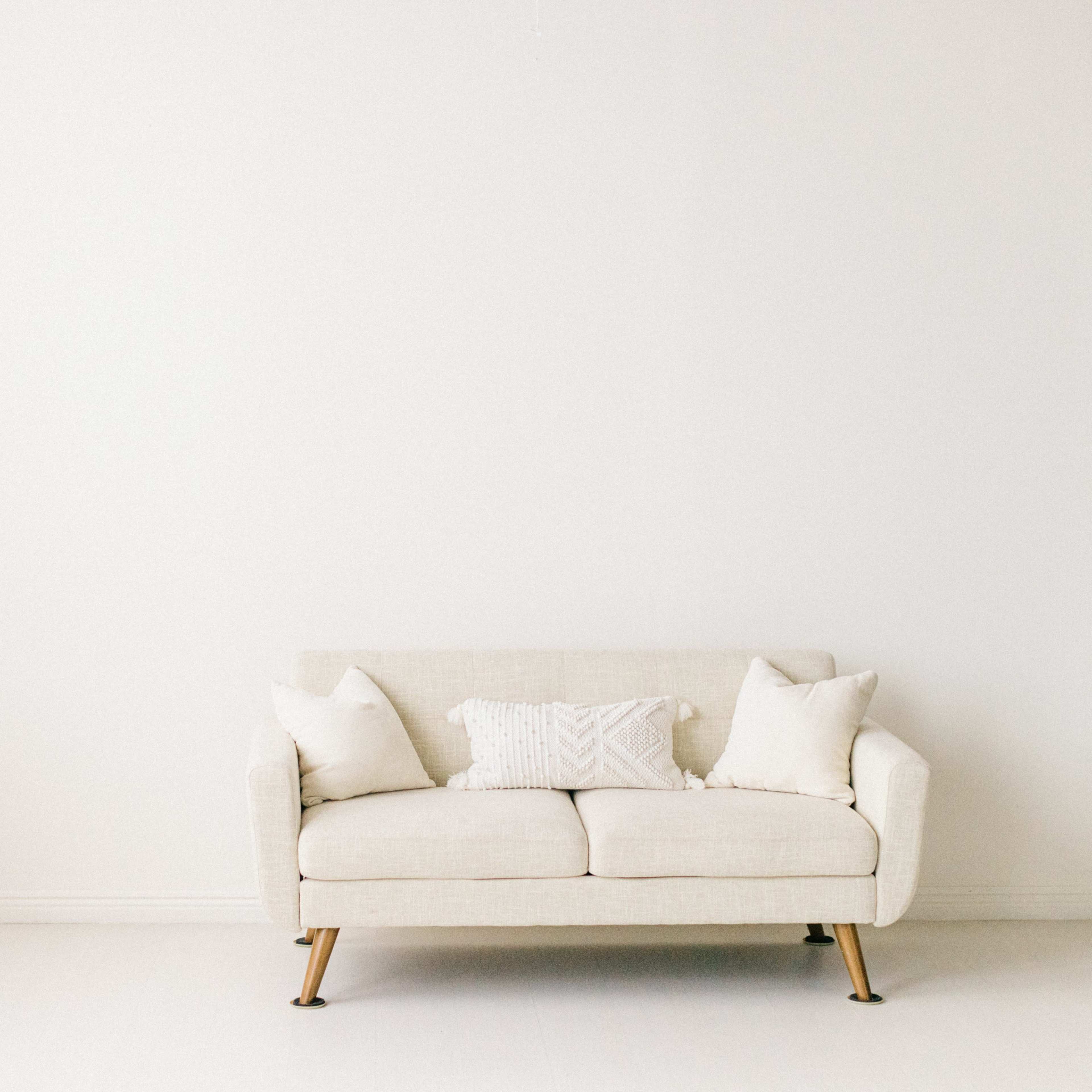A light-colored sofa with two cushions sits against a plain white wall in a bright, minimalistic room.