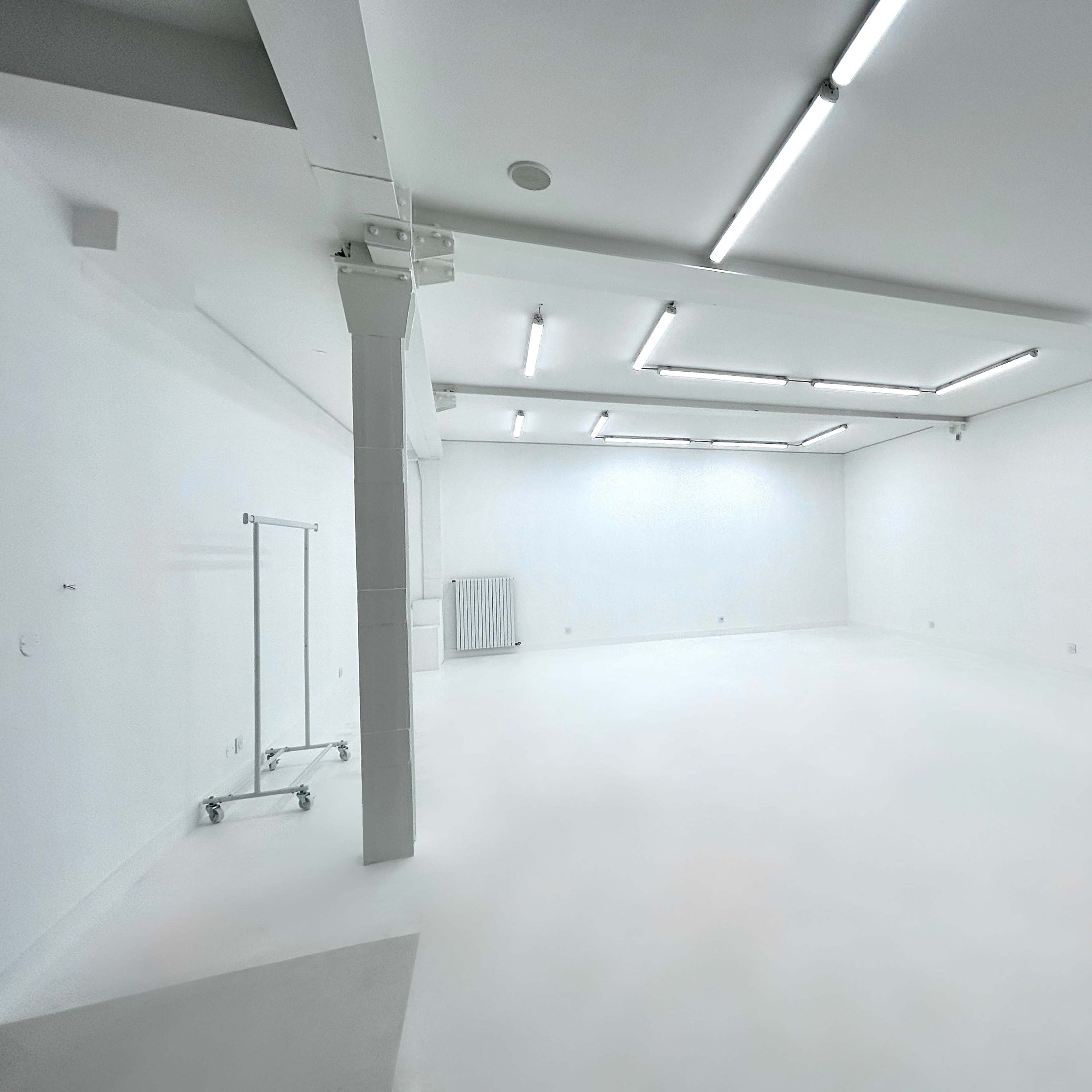 A spacious, minimalist room with white walls and a polished floor, featuring fluorescent ceiling lights and a clothing rack in one corner.