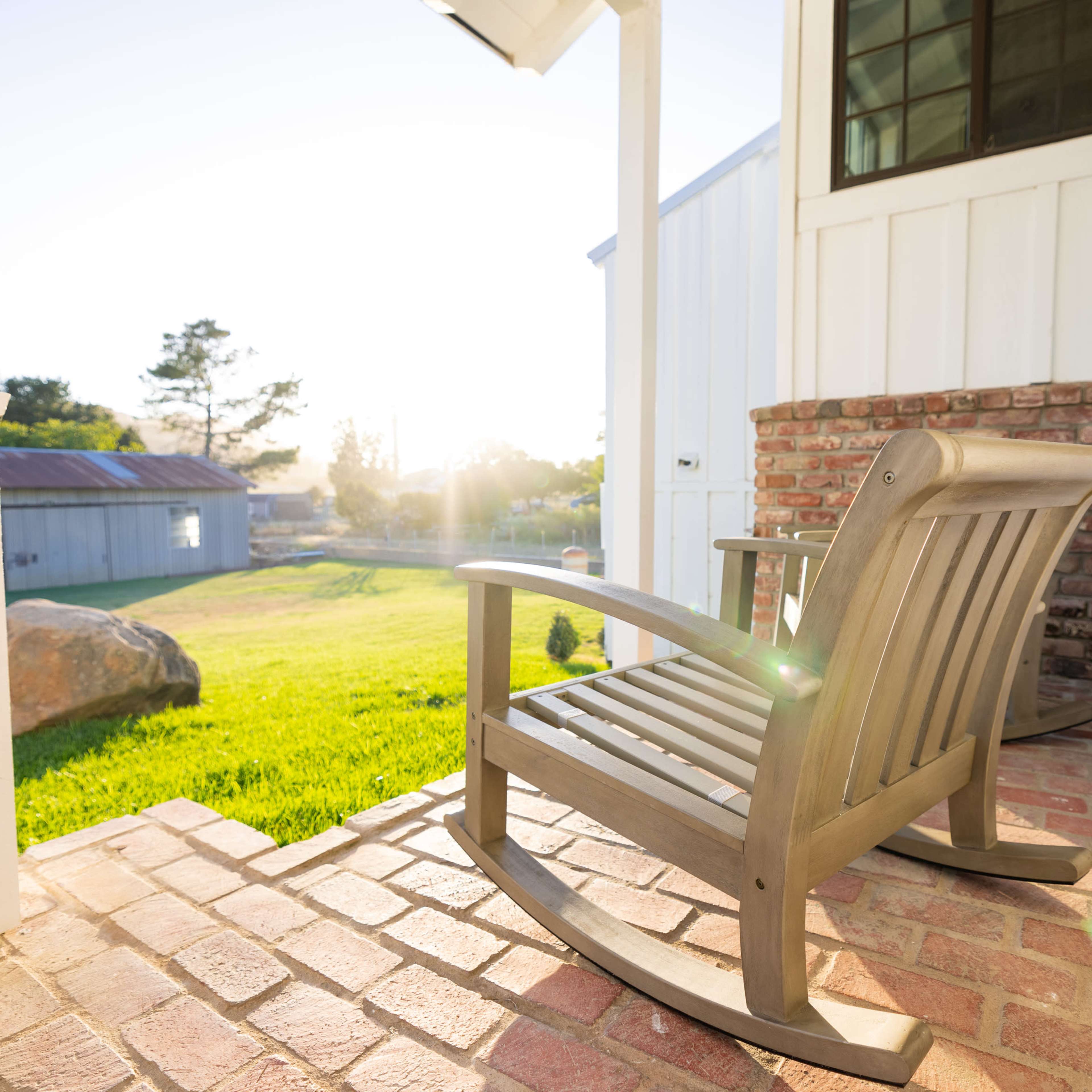 A wooden rocking chair sits on a brick porch overlooking a grassy area and a shed in the background.