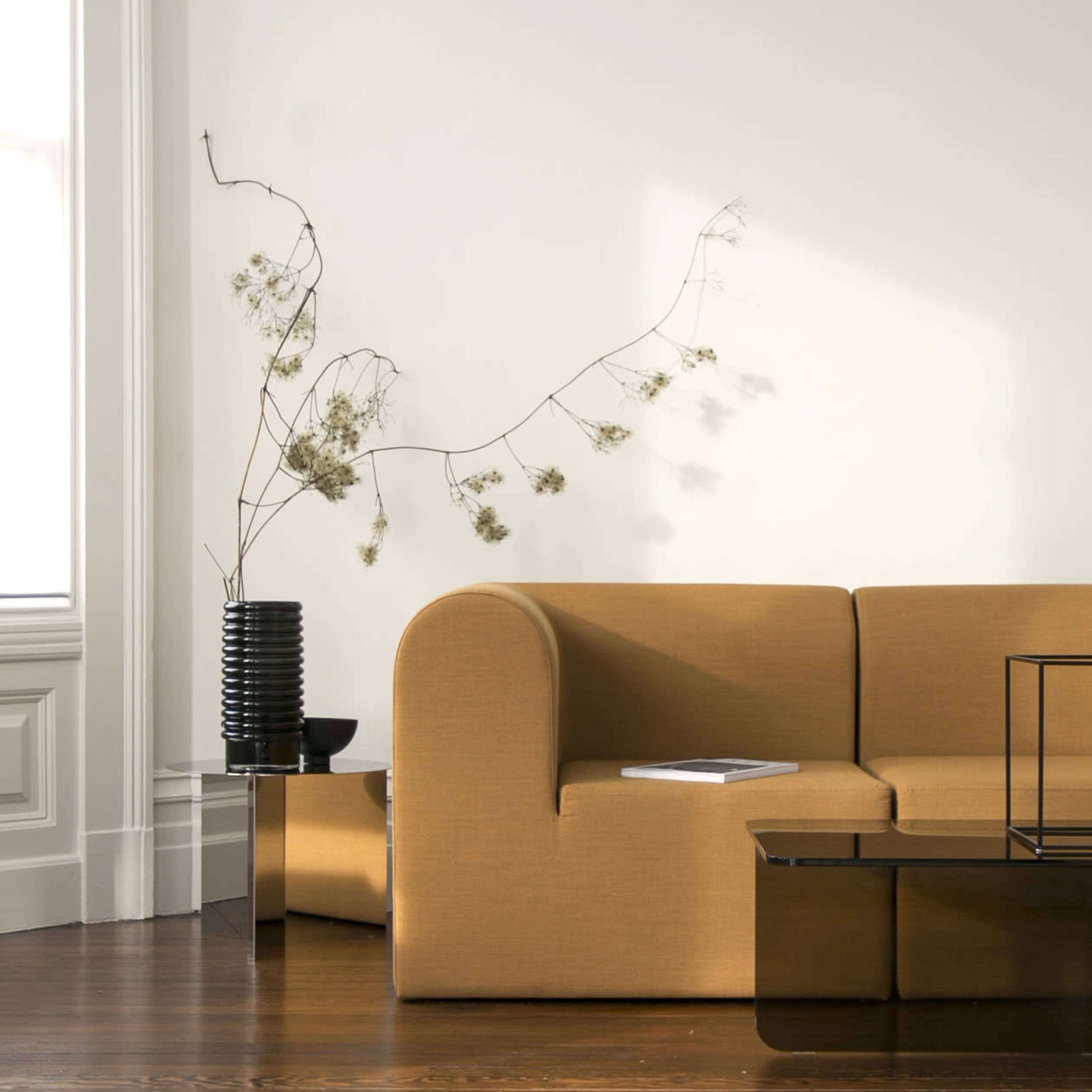 A curved yellow sofa sits against a white wall, next to a black vase with dried flowers and a coffee table.