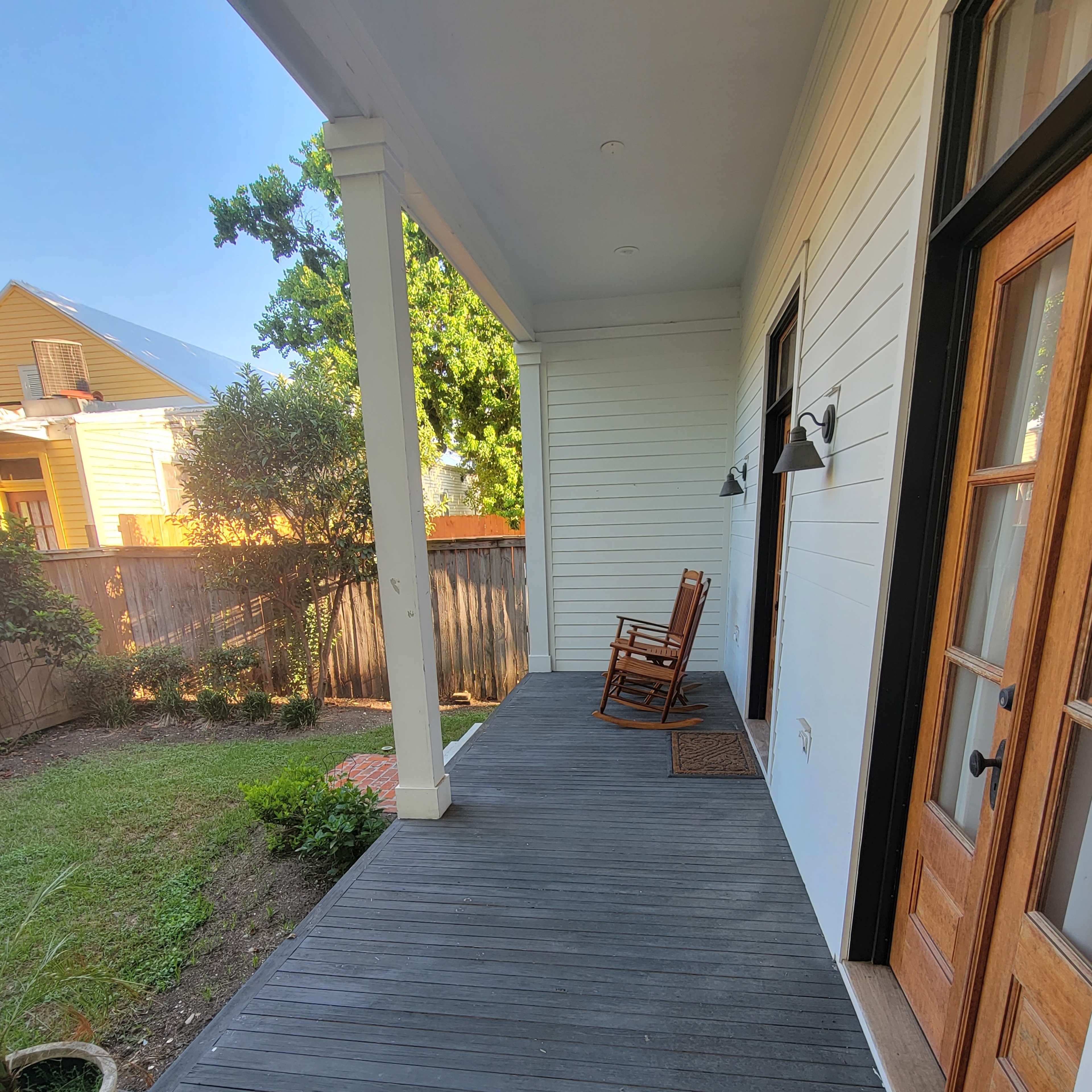 A covered porch with a rocking chair, wooden doors, and a view of a grassy yard beyond a low wooden fence.