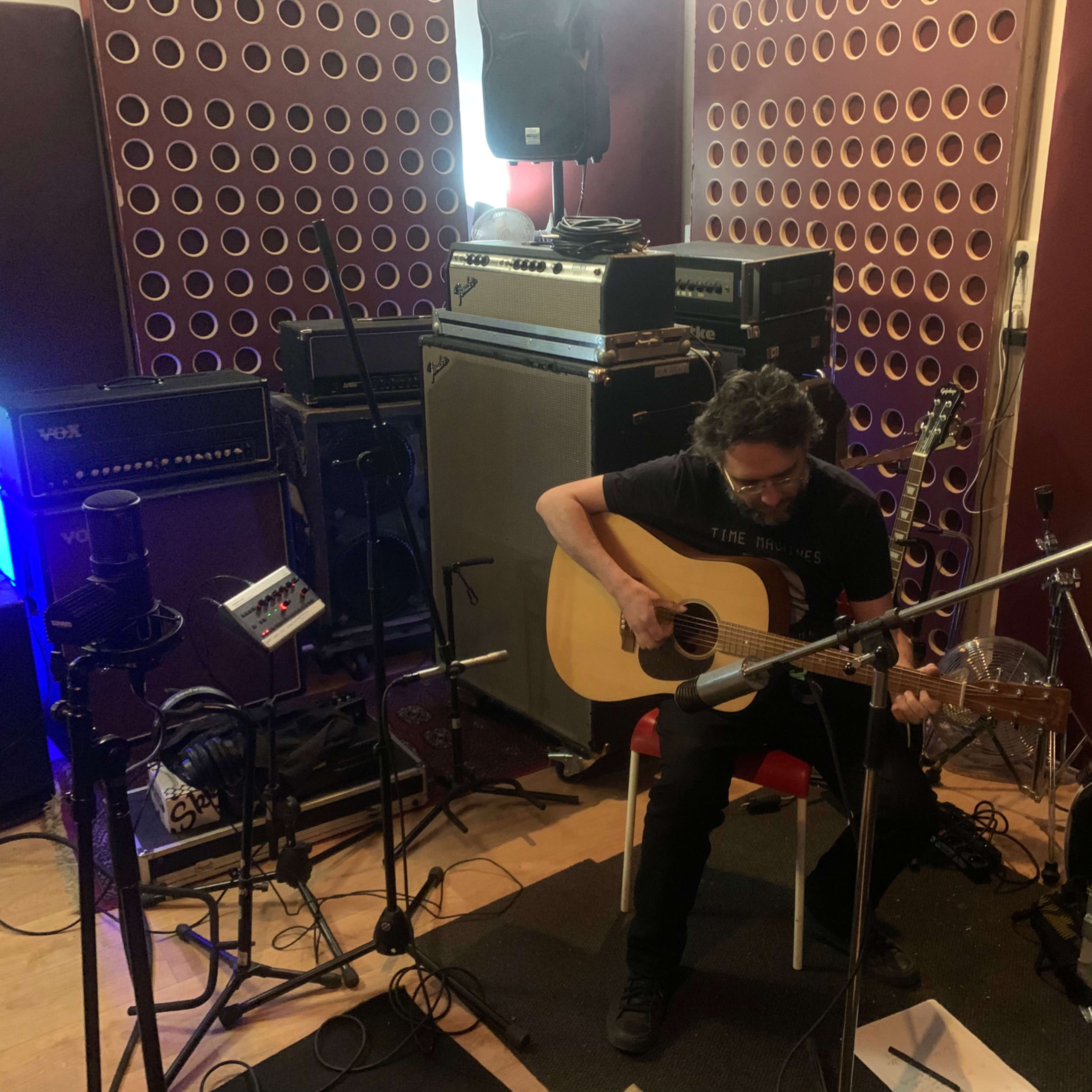 A musician sits on a chair playing an acoustic guitar in a recording studio surrounded by amplifiers and audio equipment.
