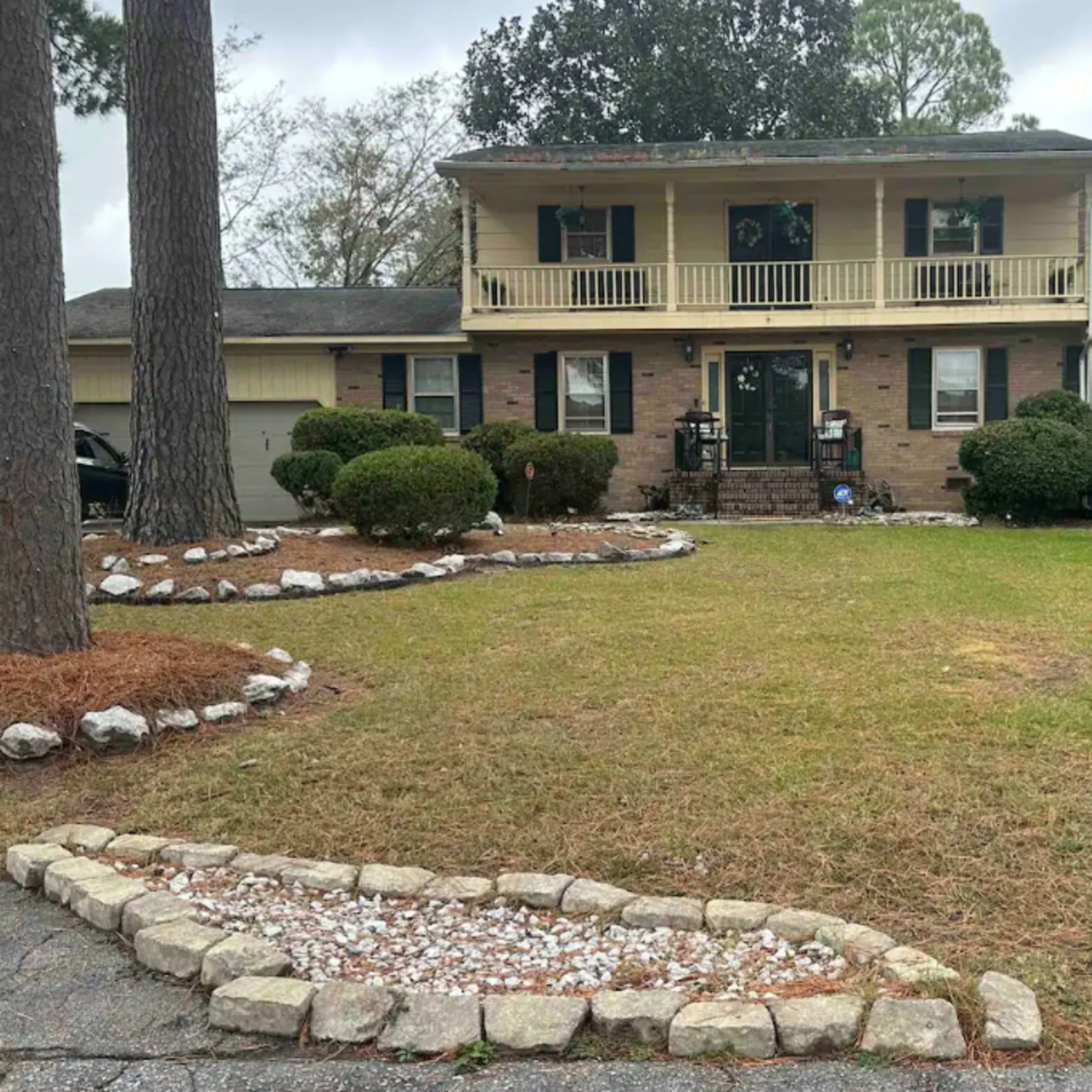A two-story house with a front porch, flanked by trees and a landscaped yard featuring a rock-bordered flower bed.