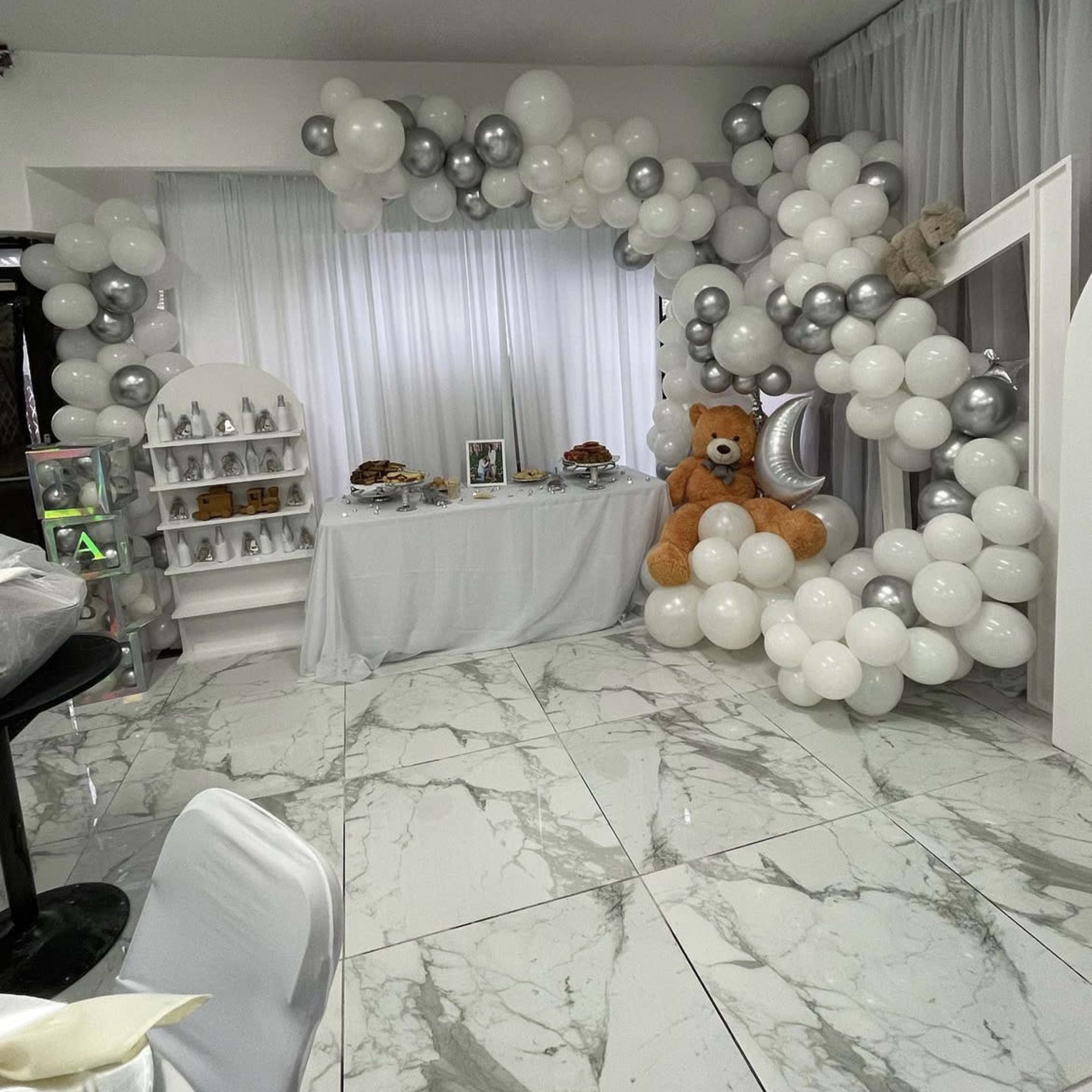 A decorated event space featuring a backdrop of white and silver balloons alongside a table with snacks and a large teddy bear.
