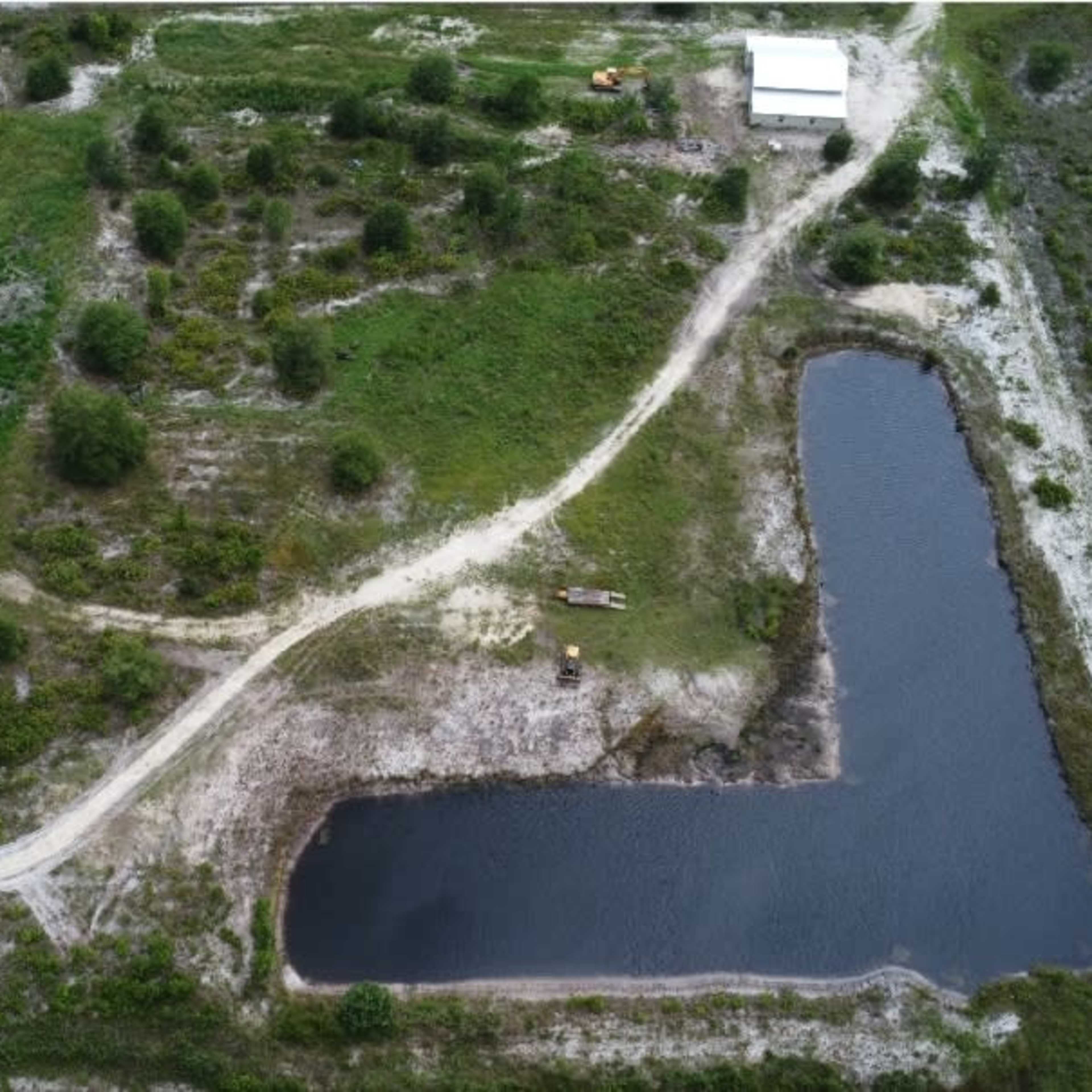 An aerial view of a large, vacant plot of land with a winding dirt path, a small pond, and some scattered trees.