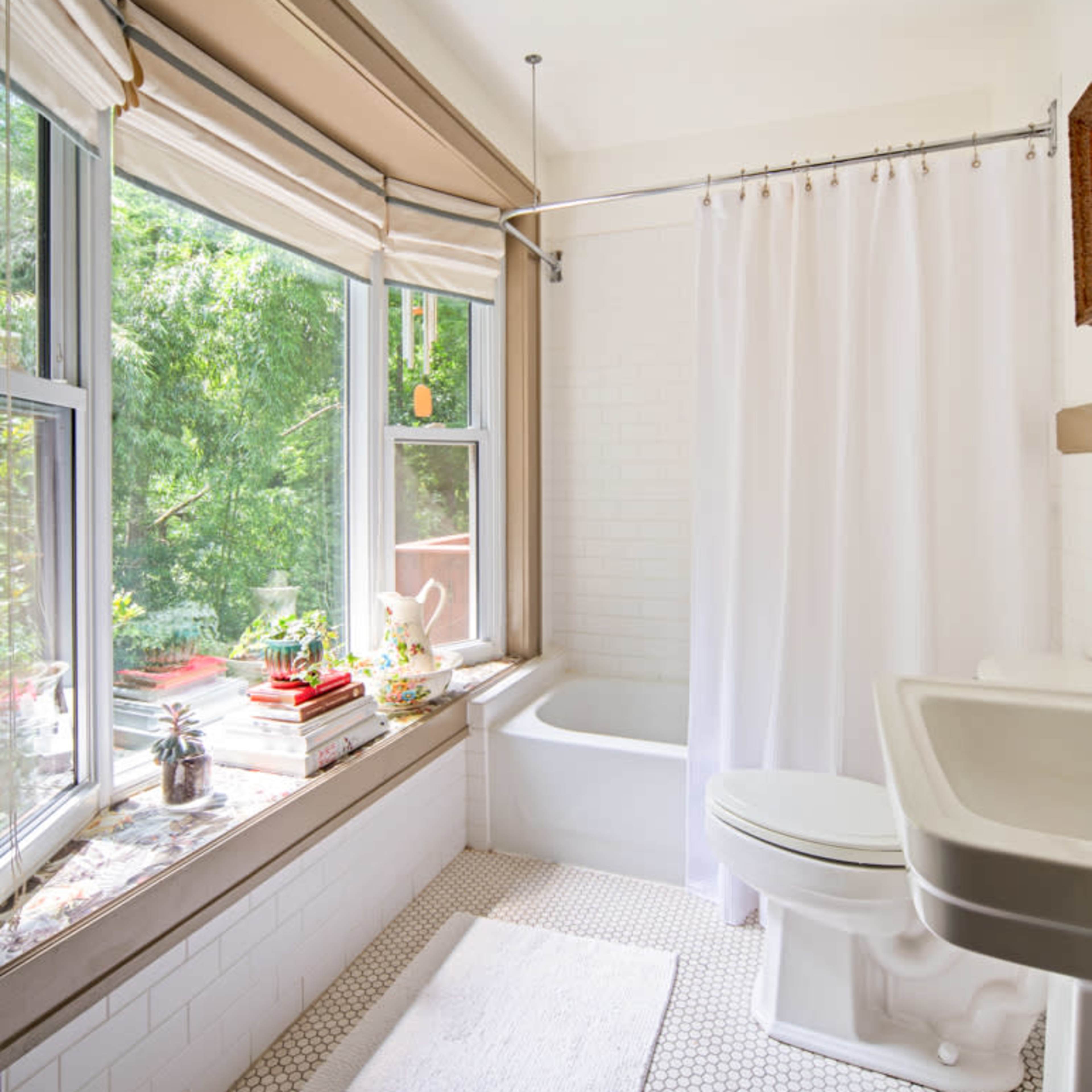 A bright bathroom with a bathtub, white shower curtain, and a window featuring lush greenery outside.