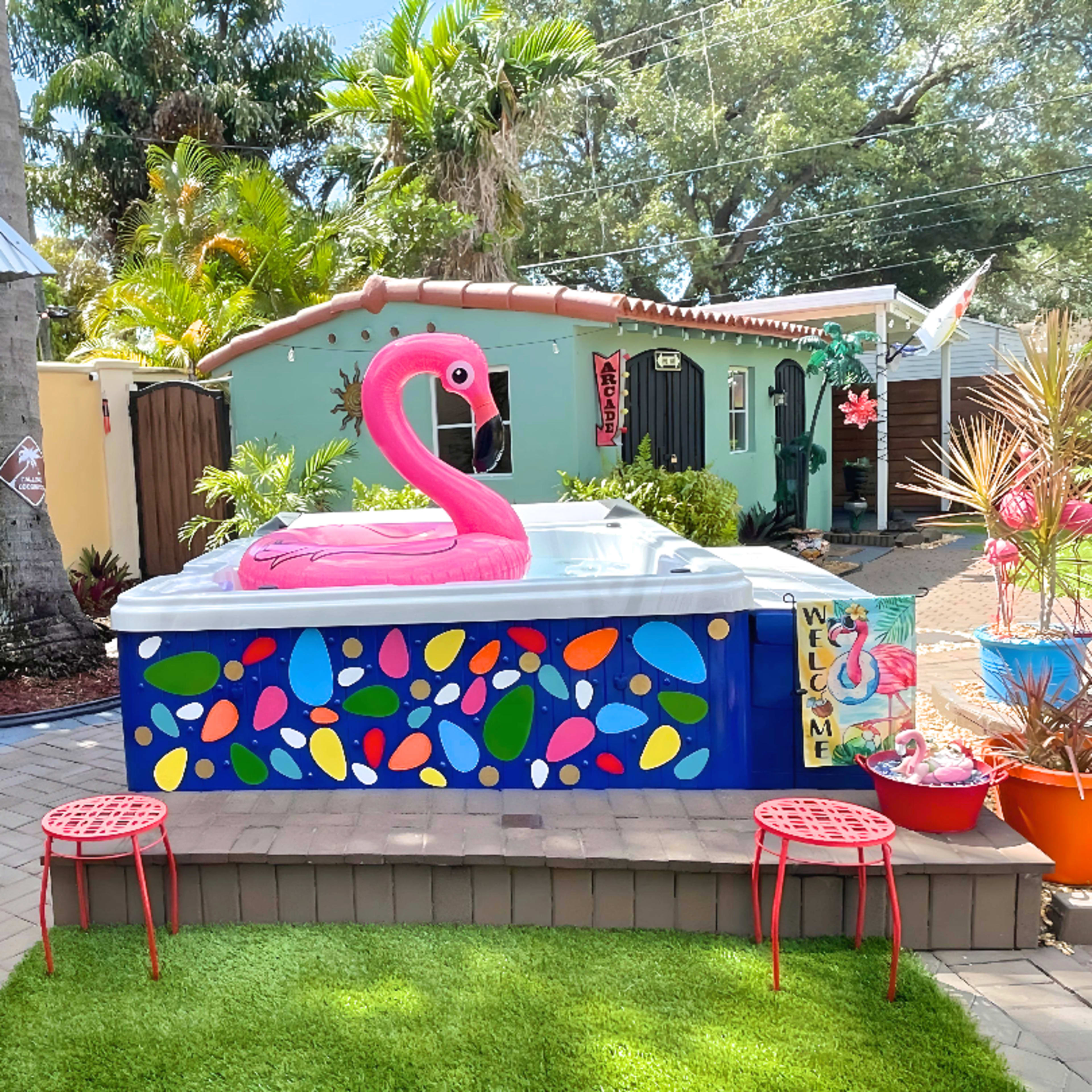 A vibrant hot tub surrounded by colorful decor and tropical plants, featuring a large flamingo float and two red chairs on green grass.