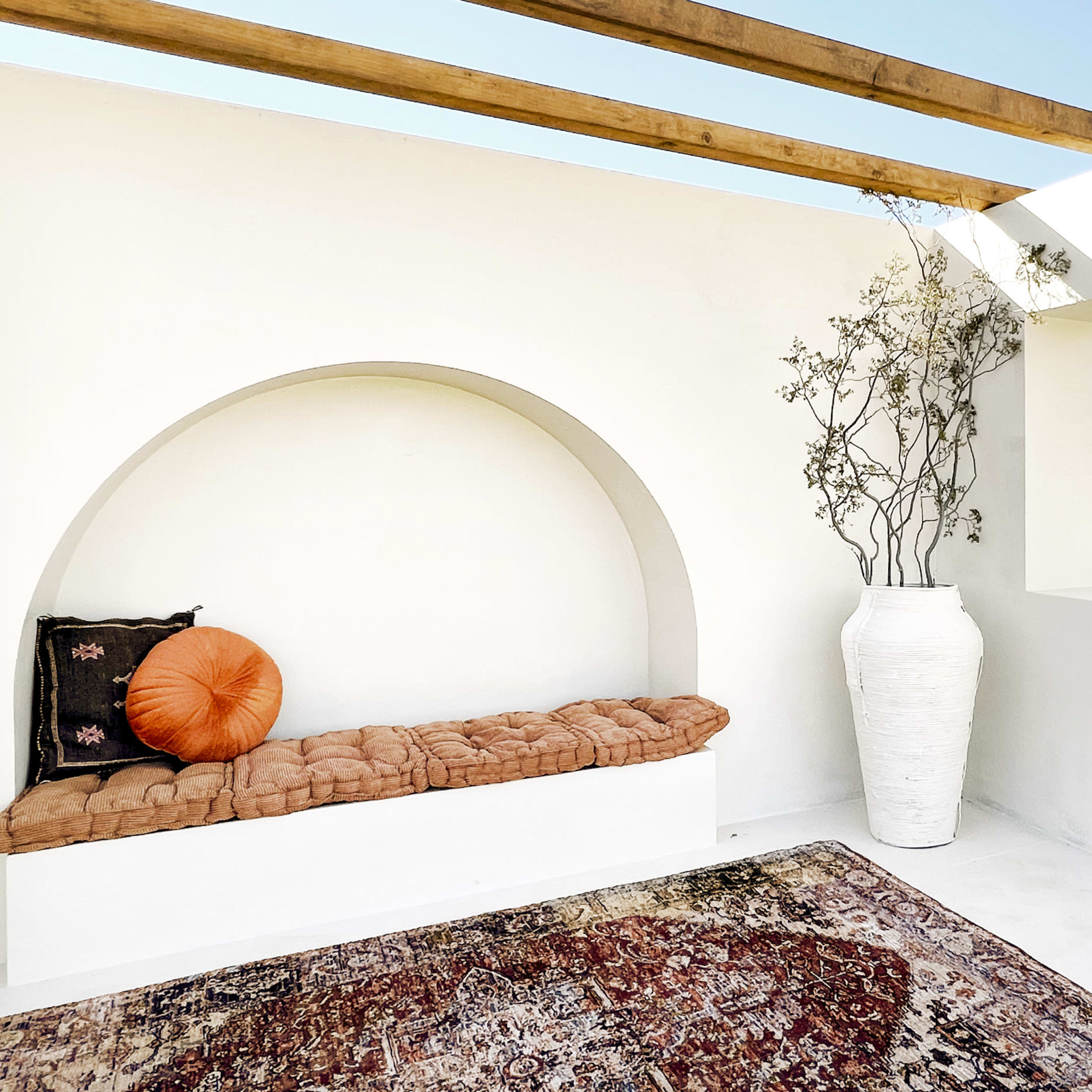 A minimalist indoor space with a cushioned seating area, a large white vase, and an opening that frames a view of the outdoors.