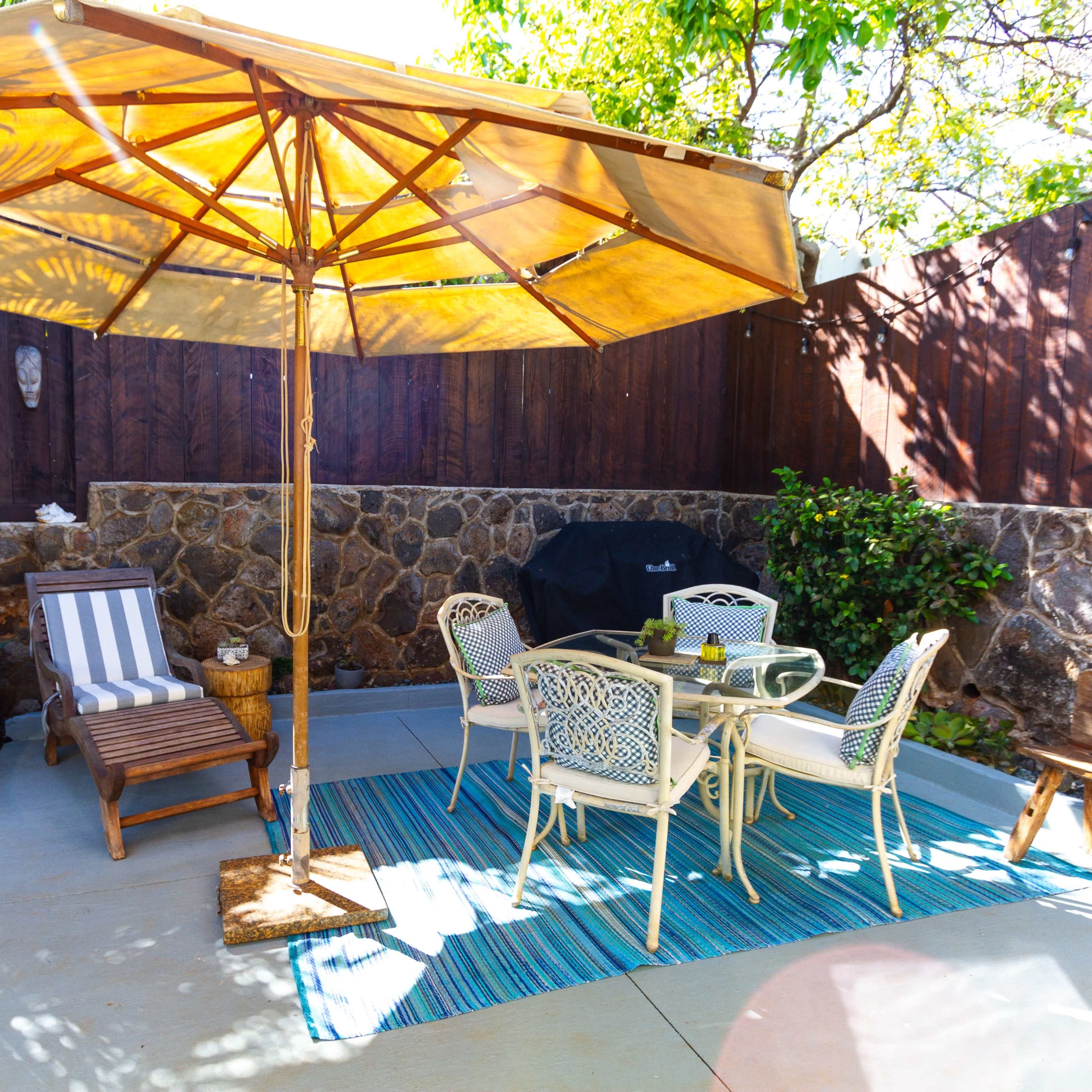 A patio area features a tan umbrella shading a table with four chairs, accompanied by a lounge chair and a grill against a stone wall.