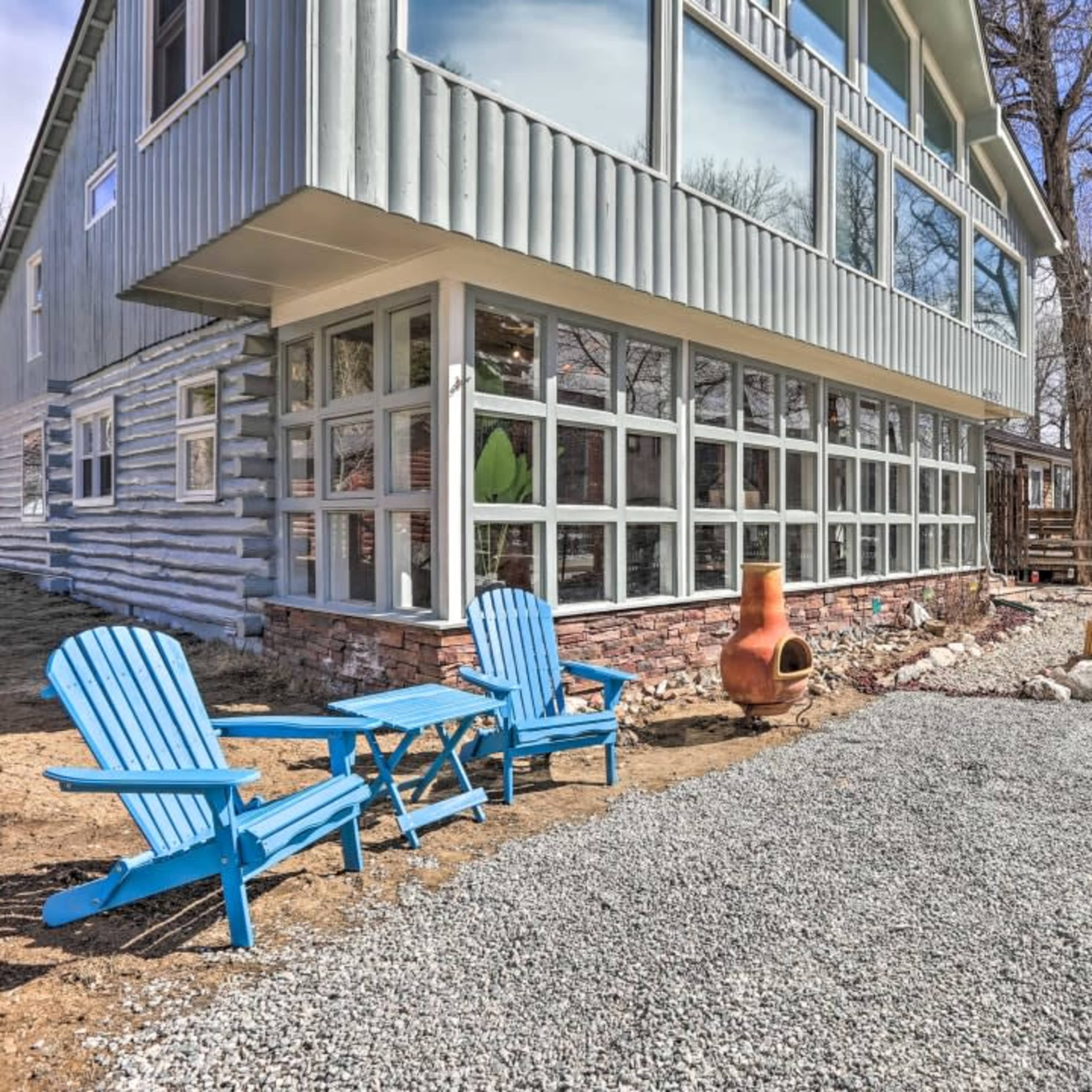 A home with a large glass wall features two blue adirondack chairs and a gravel area near a chimney in a yard surrounded by trees.
