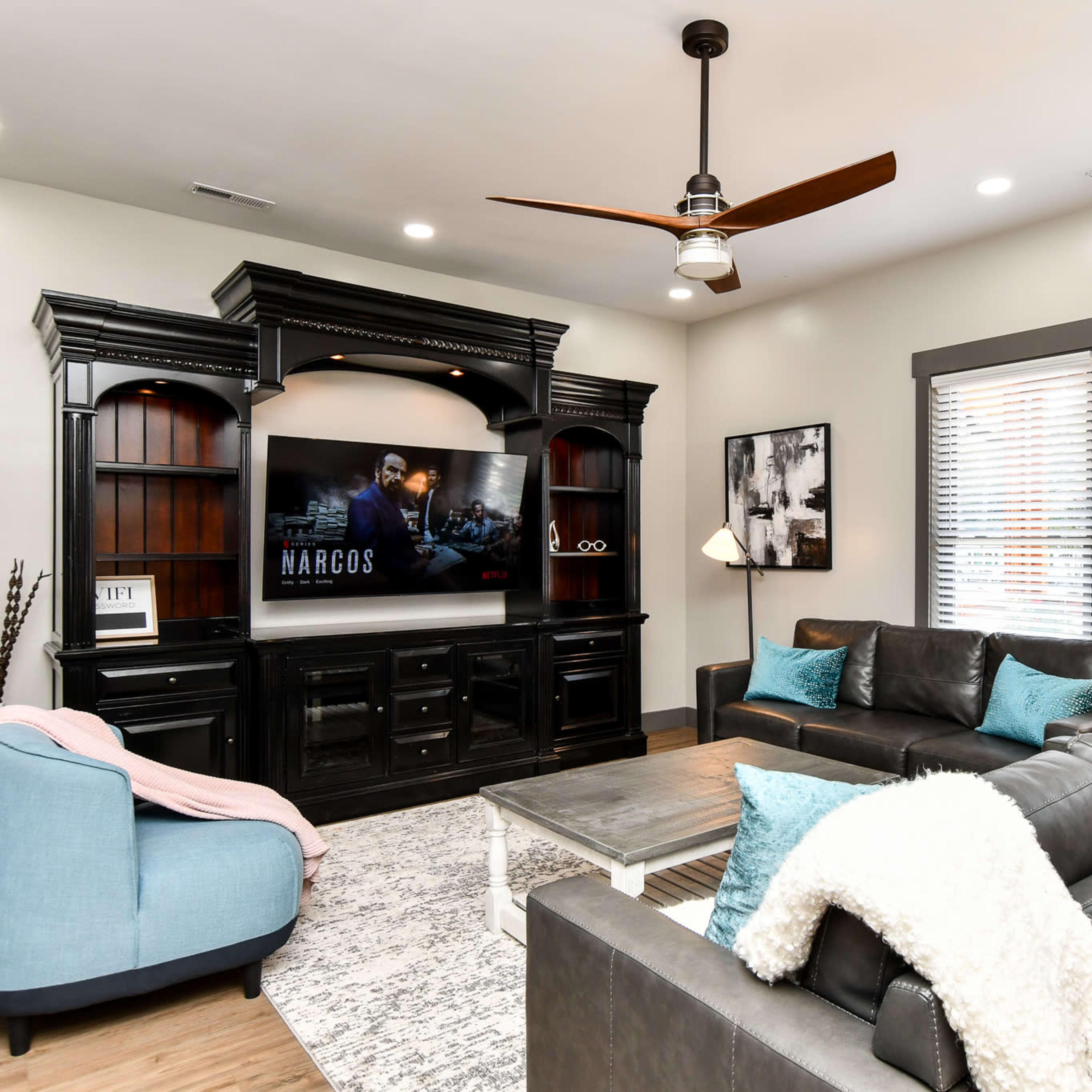 The living room features a dark wooden entertainment center with a mounted TV displaying a show, a black leather sofa with decorative pillows, and a light-colored area rug.