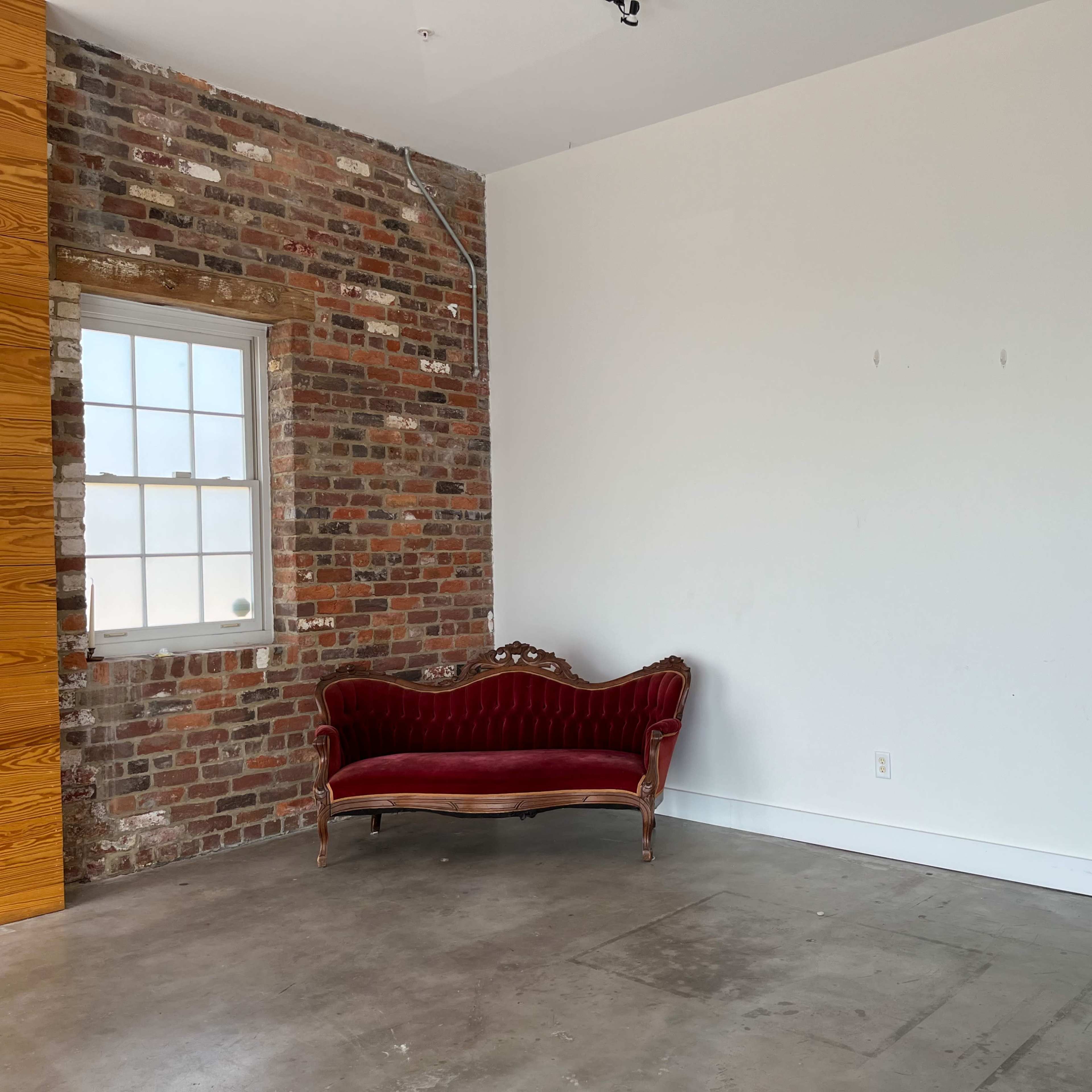 A room with a brick wall on one side, featuring a red velvet sofa and a white office chair on a concrete floor.