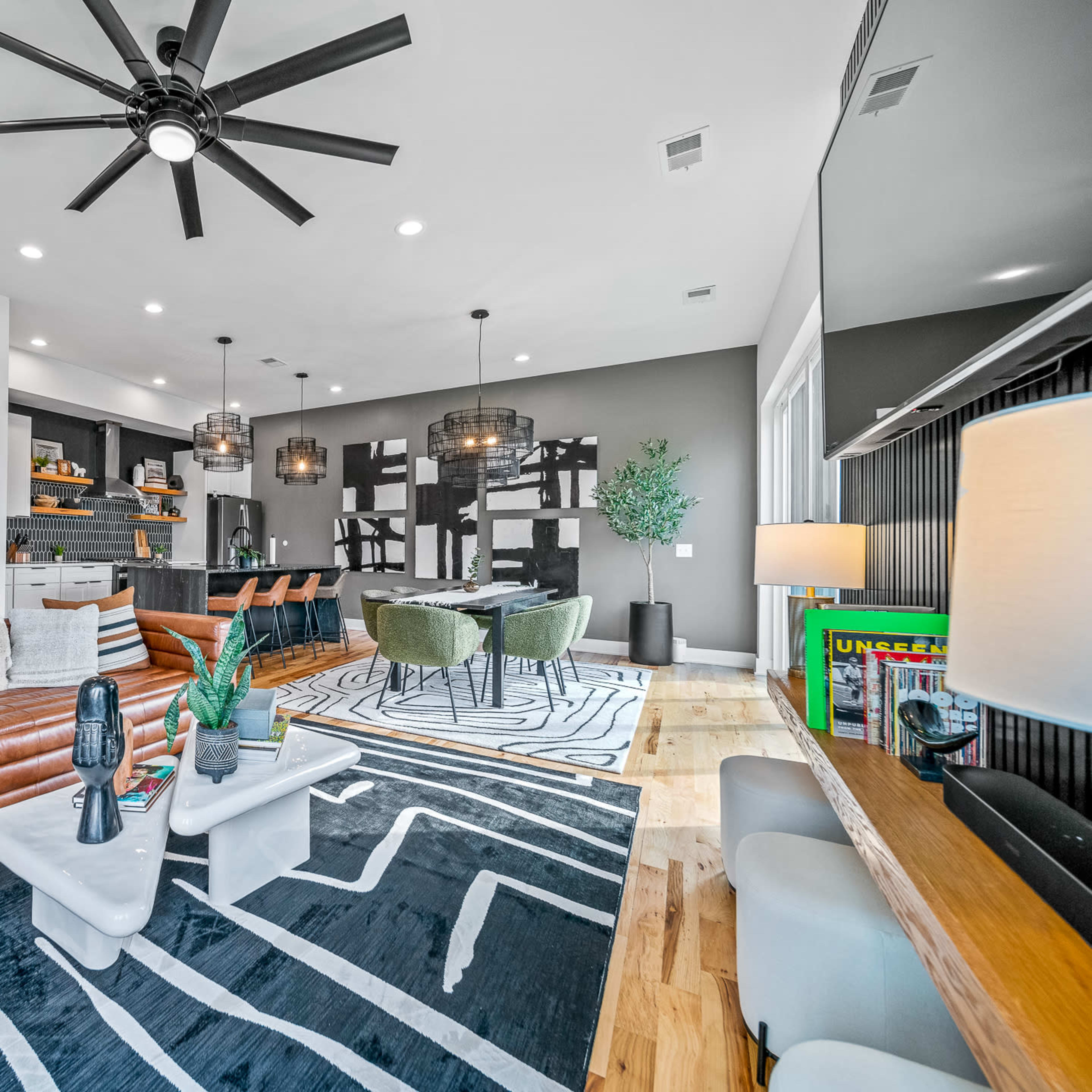 A modern living room features a leather sofa, a striped area rug, and a kitchen area with dark cabinetry and pendant lighting.